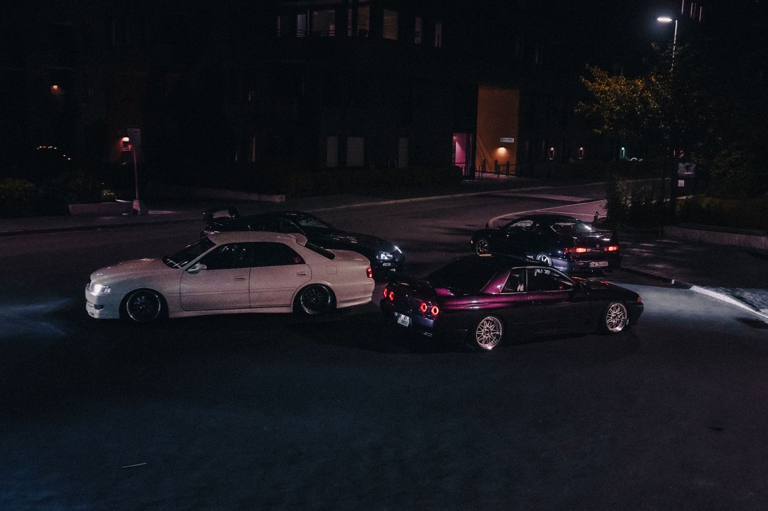 Three Japanese sports cars parked on a dark street at night, including a white and purple sedan.