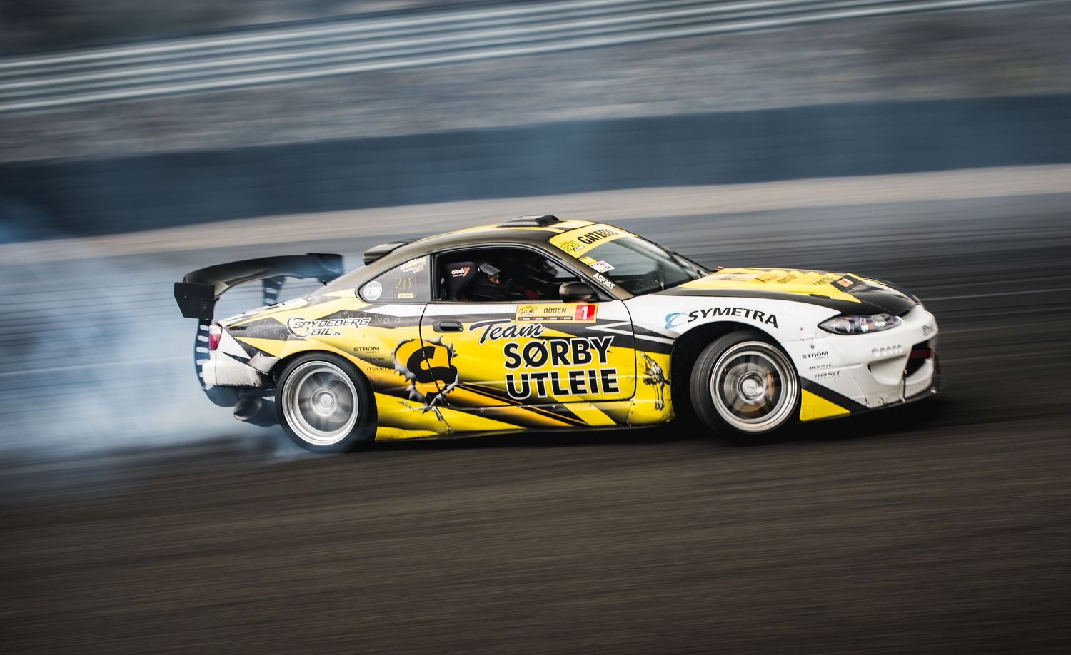 Yellow and white Nissan Silvia drift car with sponsor decals sliding on a track.