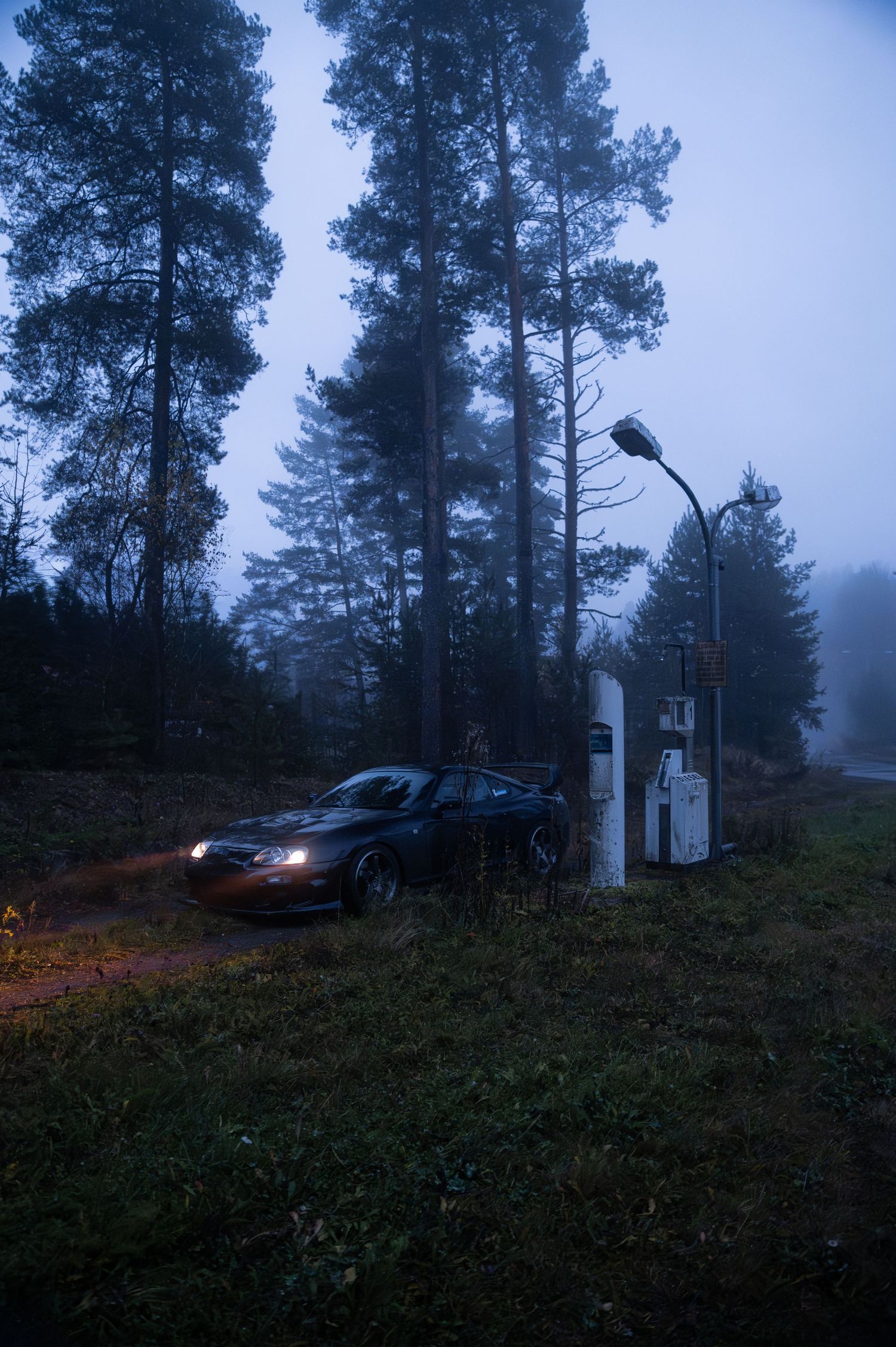 Dark sports car with headlights on at an abandoned foggy forest gas station at dusk.