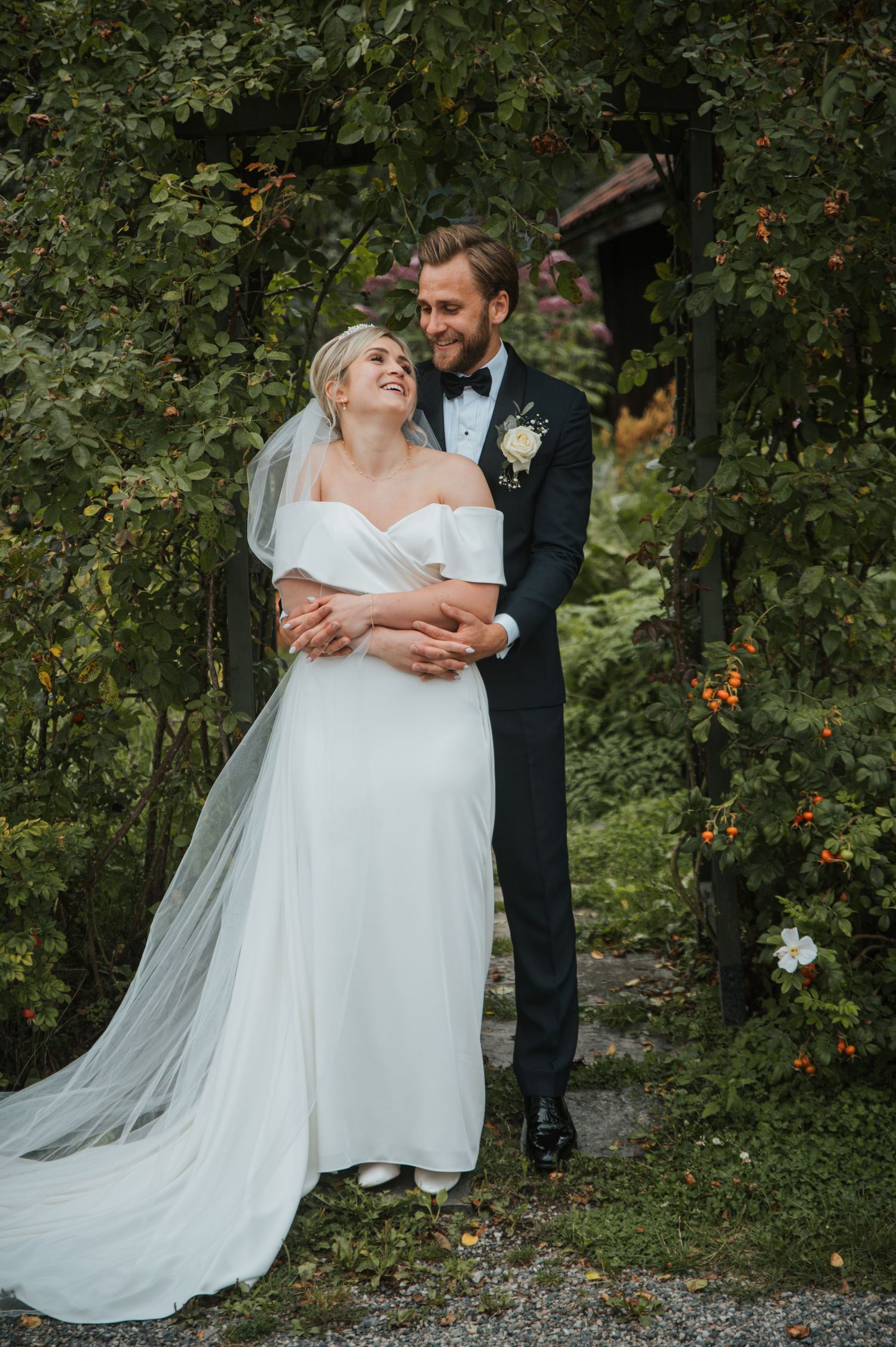 Smiling bride in off-shoulder white gown with veil embraced by groom in dark tuxedo among lush greenery.