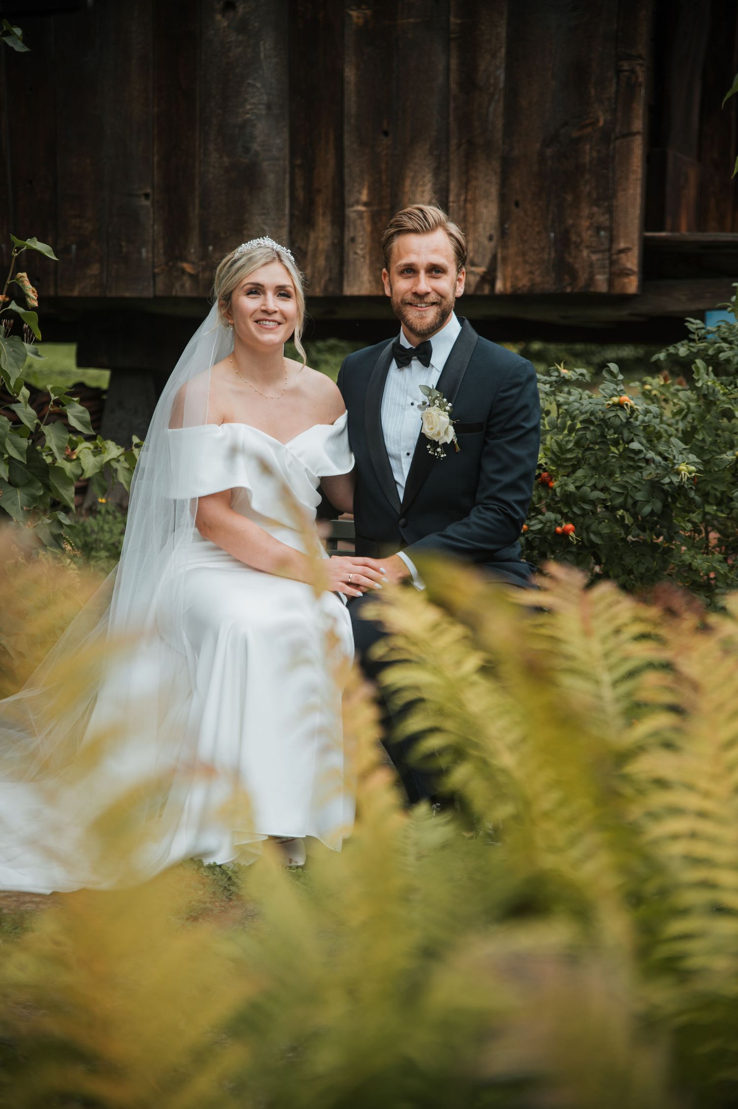 Bride in elegant off-shoulder white dress and groom in dark suit seated together among ferns and foliage.