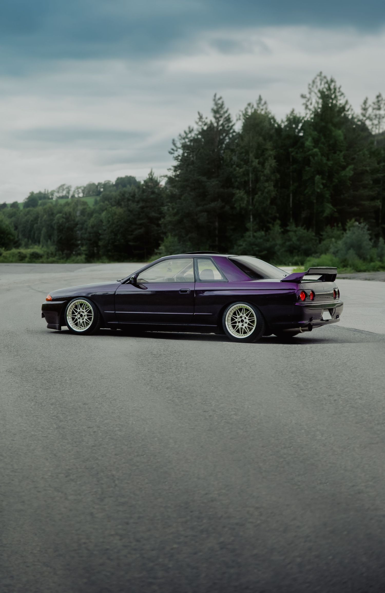 Purple Nissan Skyline R32 GT-R side profile with BBS wheels on an open tarmac with forest backdrop.