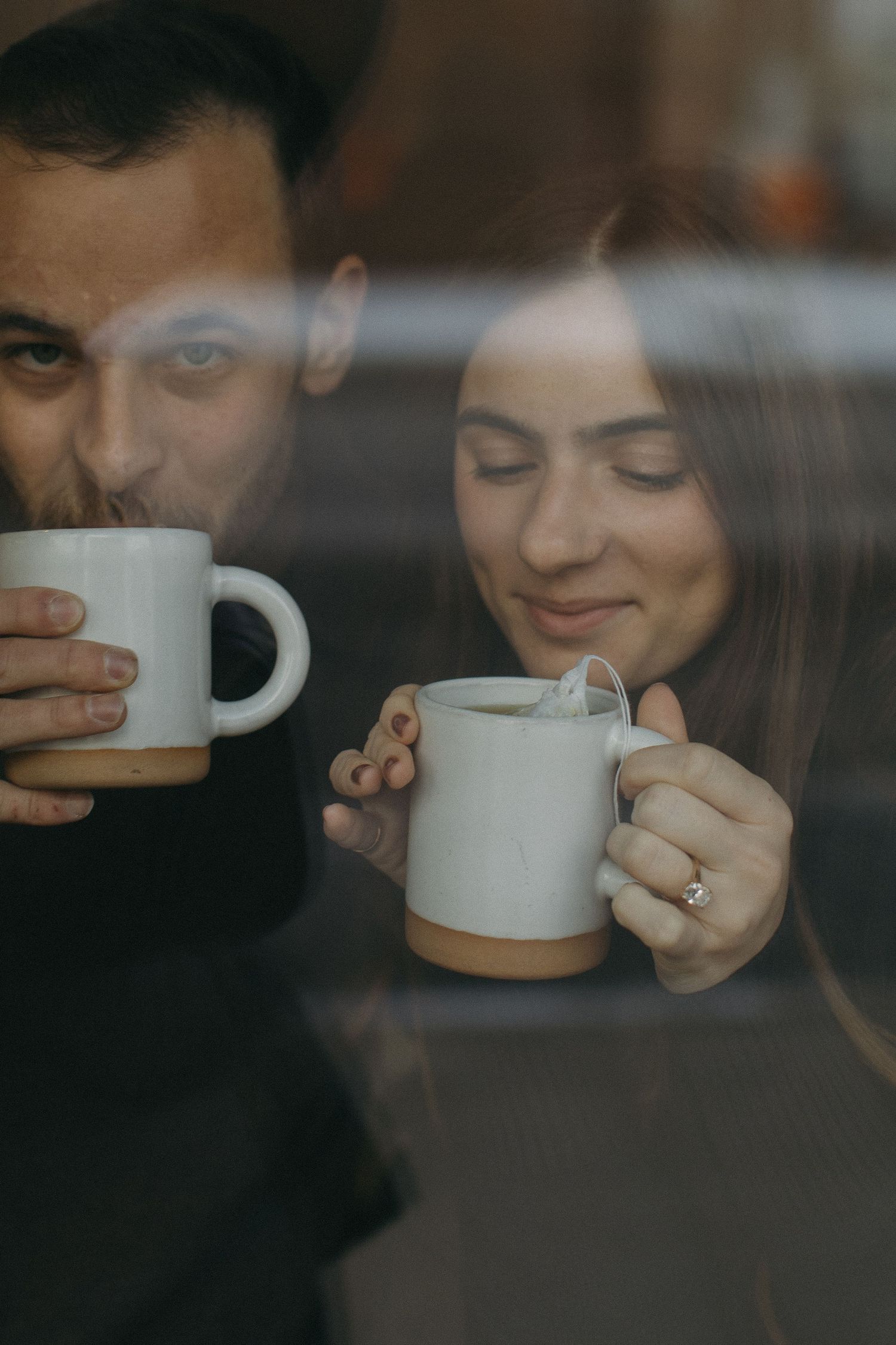 Intimate coffee date captured through window glass with coffee mugs and warm lighting.