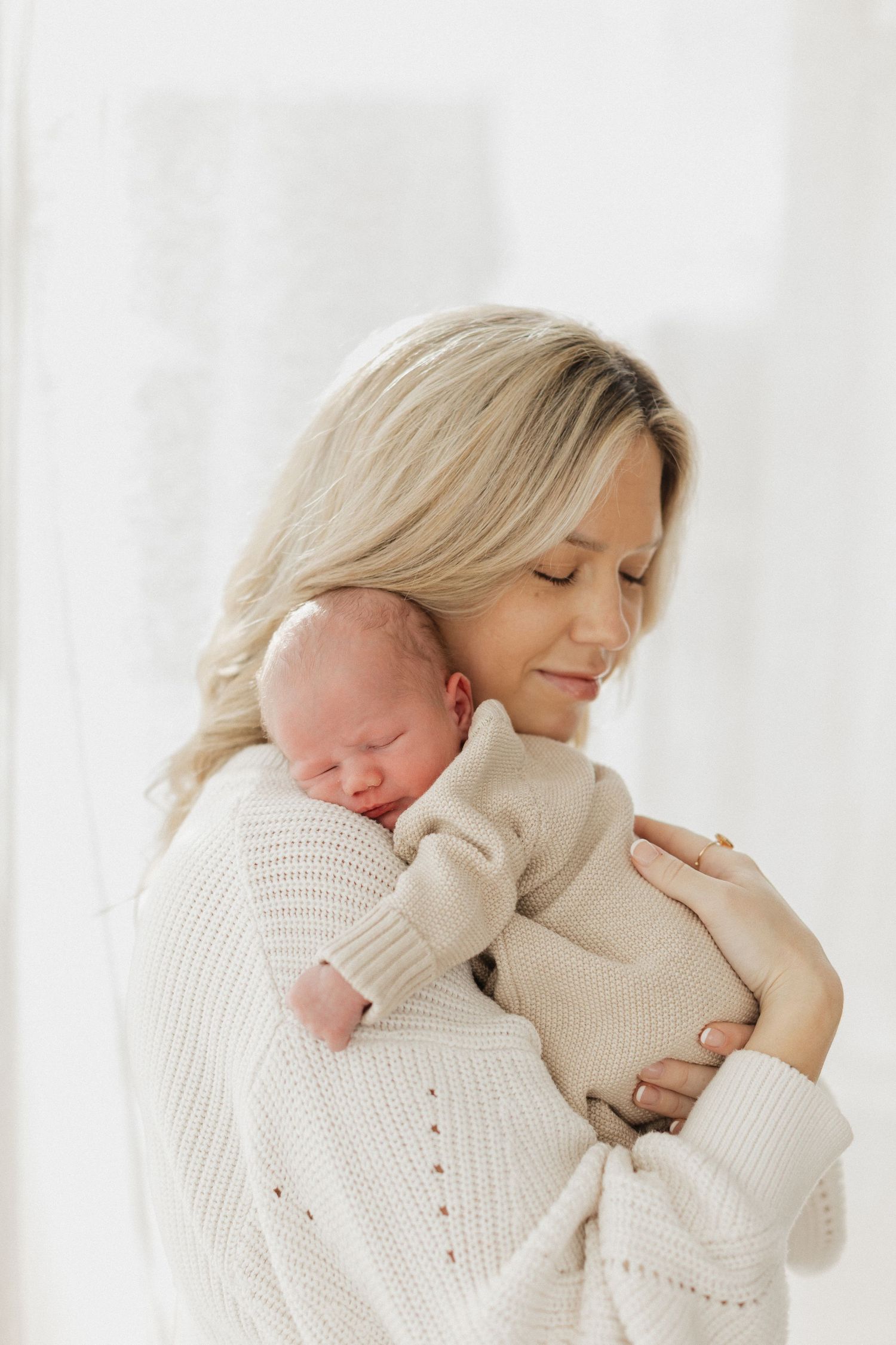 Intimate indoor portrait of mother in cream sweater holding sleeping newborn against white background.