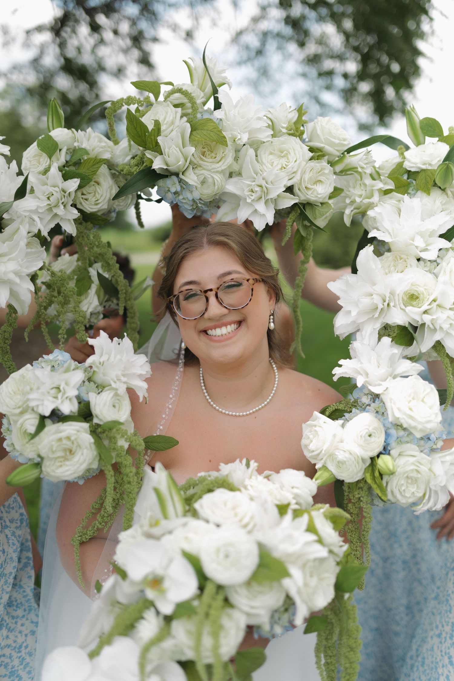 Smiling bride in glasses framed by white floral arch with roses and greenery on her wedding day.