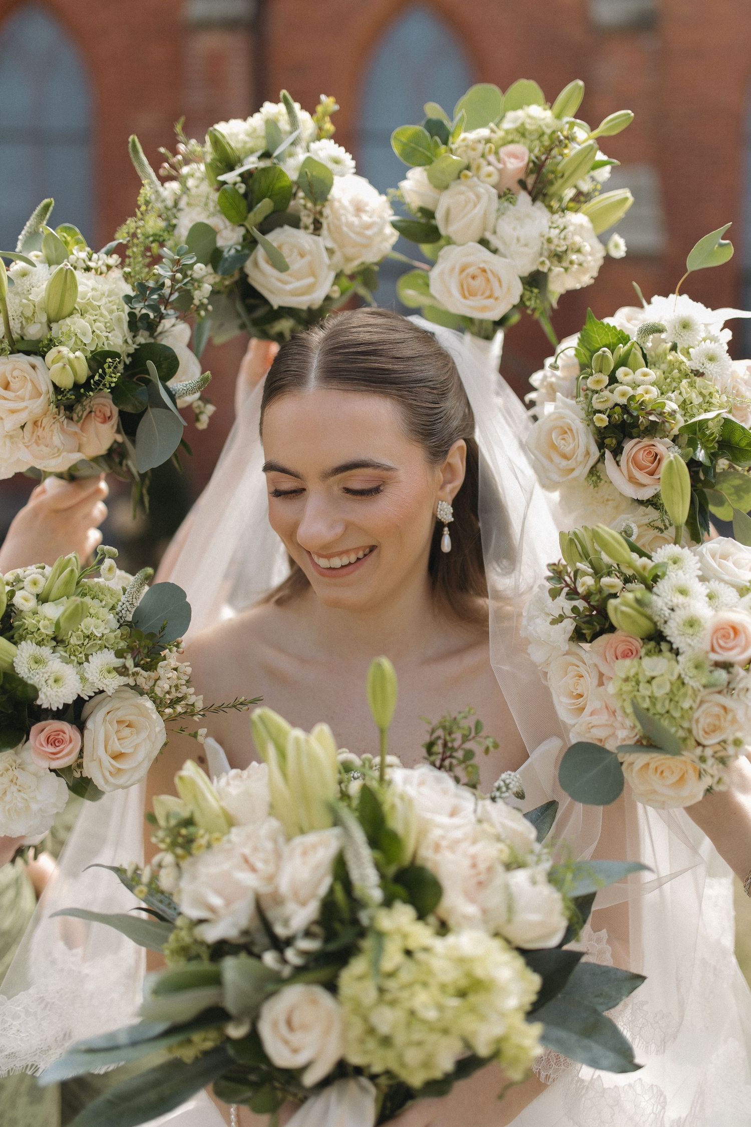 Smiling bride in white veil surrounded by elegant bouquets of roses, lilies, and greenery on her wedding day.