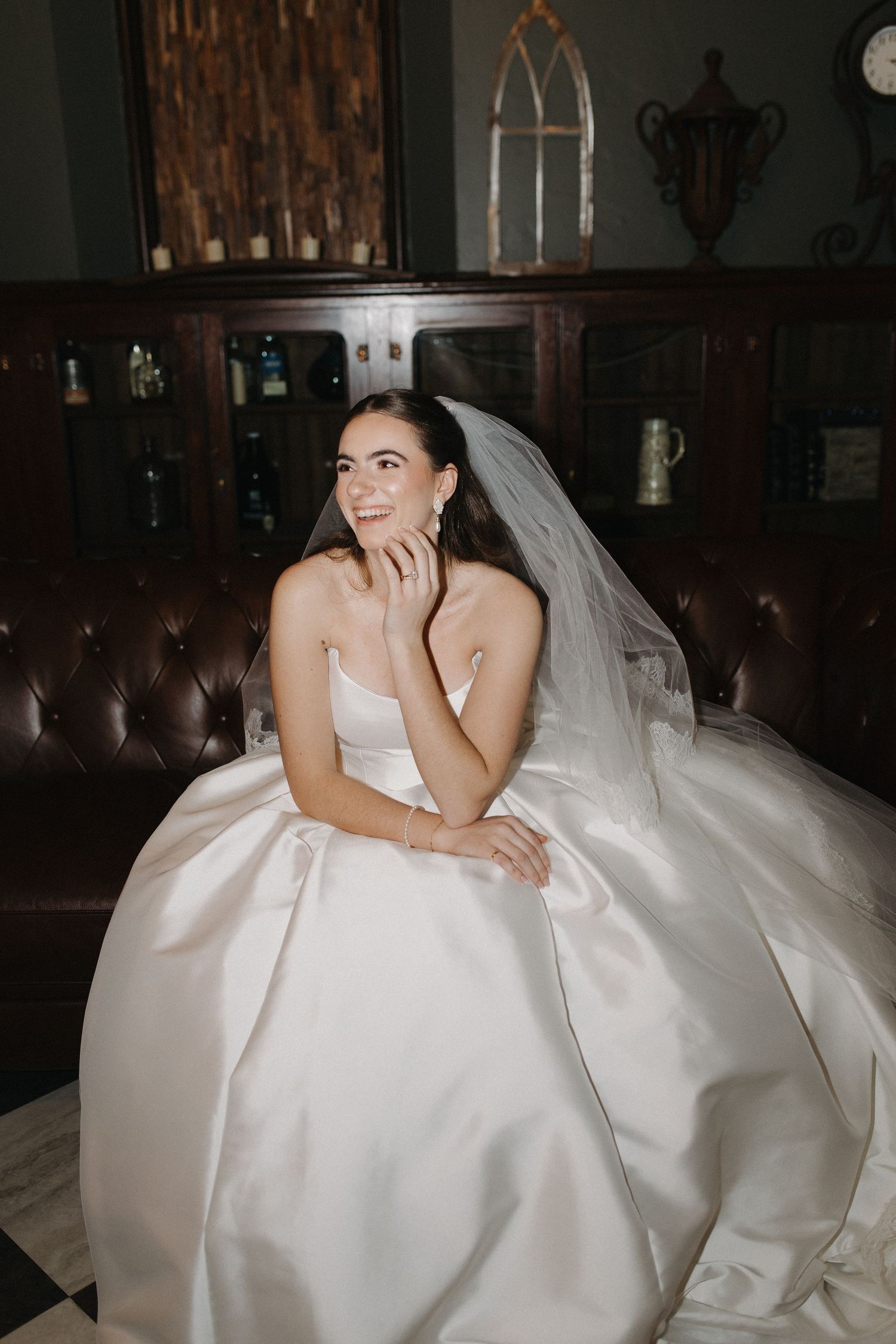 A bride in a voluminous white wedding gown sits gracefully on a dark couch in an elegant indoor setting.