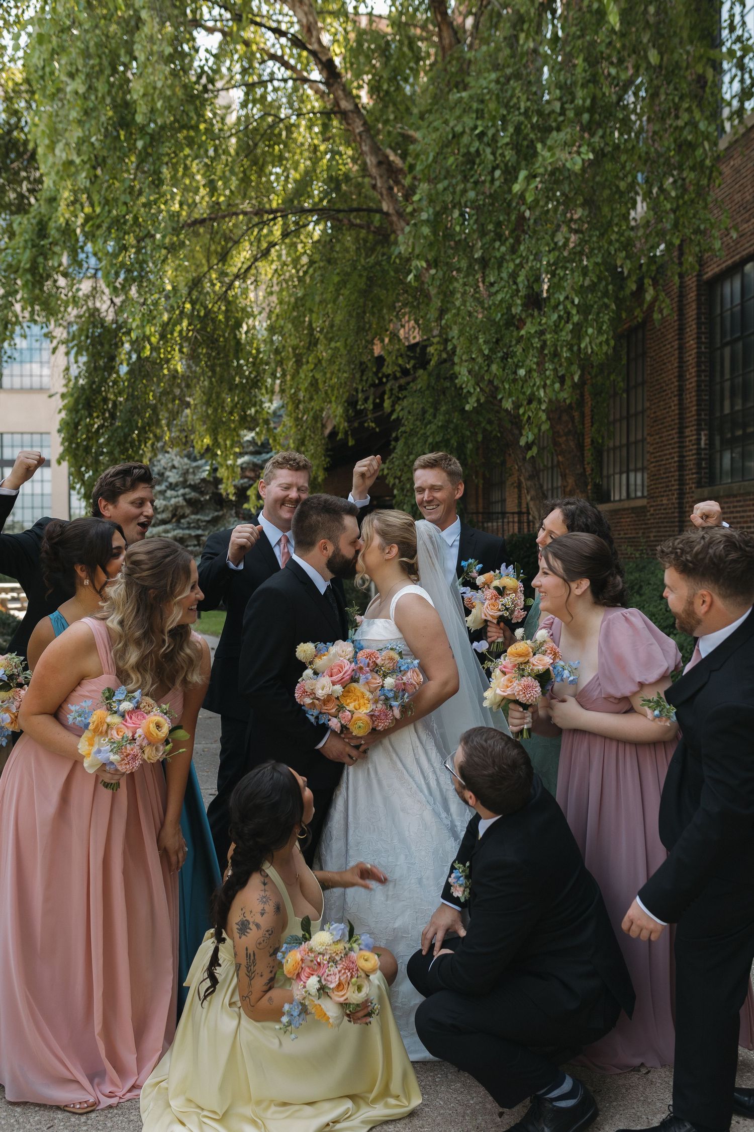 Wedding party celebrates with bride and groom outdoors, bridesmaids in pink holding colorful bouquets under a willow tree.