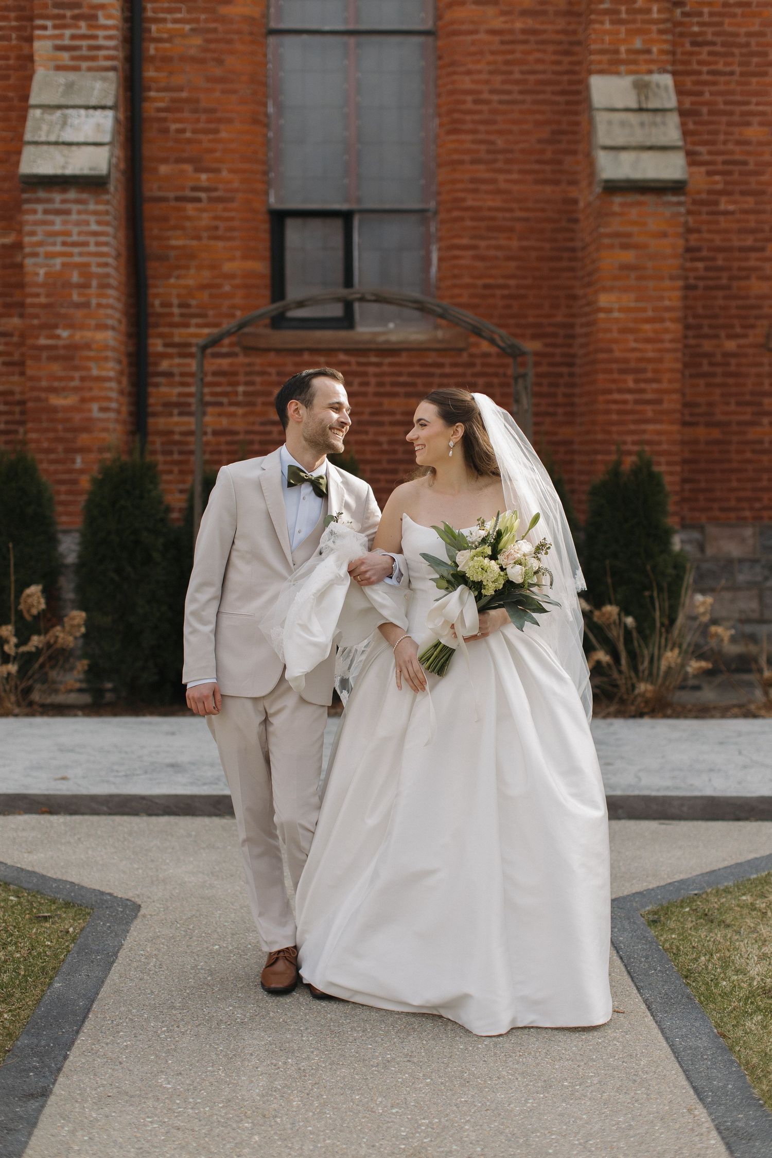 Bride and groom walking together outside a brick building, bride holding a white and yellow bouquet, groom in a tan suit.