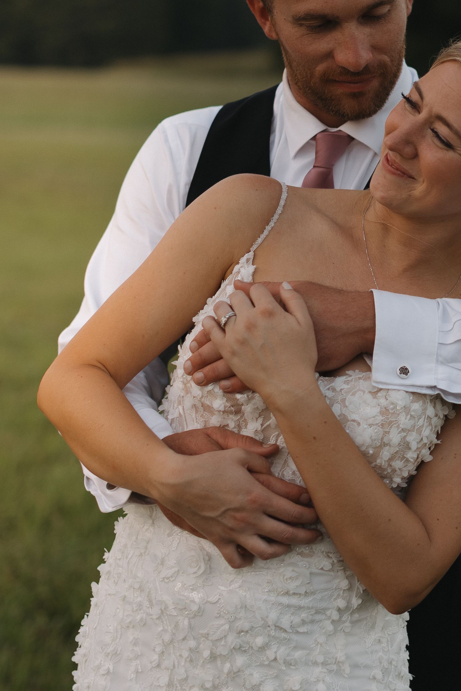 A bride and groom share a tender embrace outdoors, her white textured gown and diamond ring visible as he holds her close.
