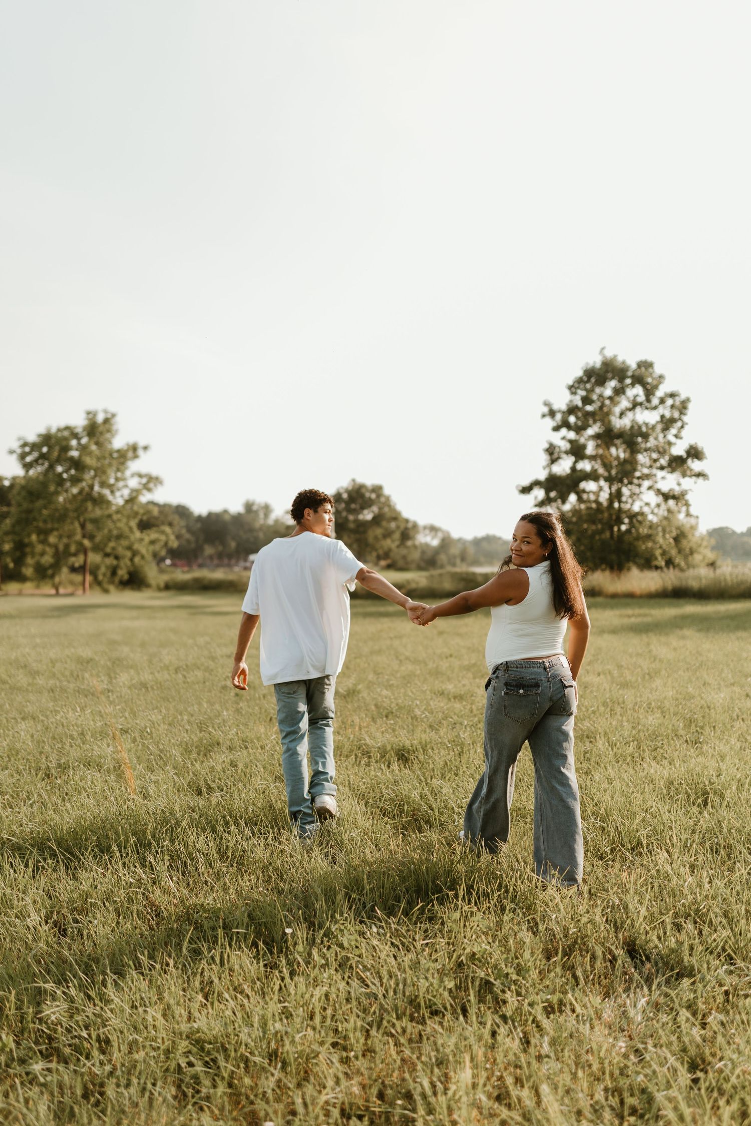A couple walks hand in hand through a grassy meadow during golden hour.