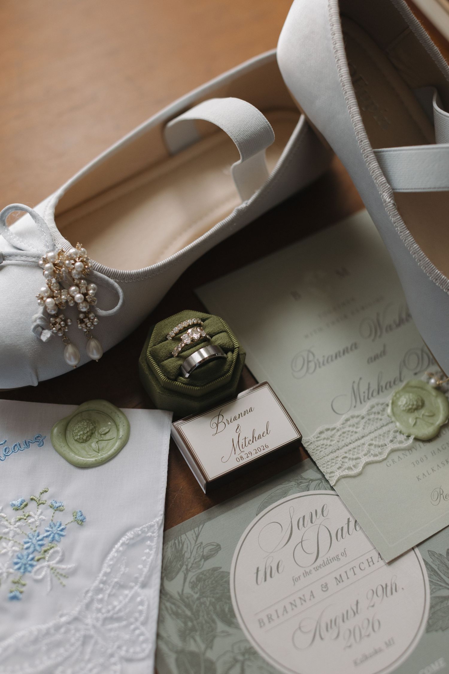 Elegant wedding flat shoes with pearl details beside green velvet ring box, save the date cards, and embroidered handkerchief.