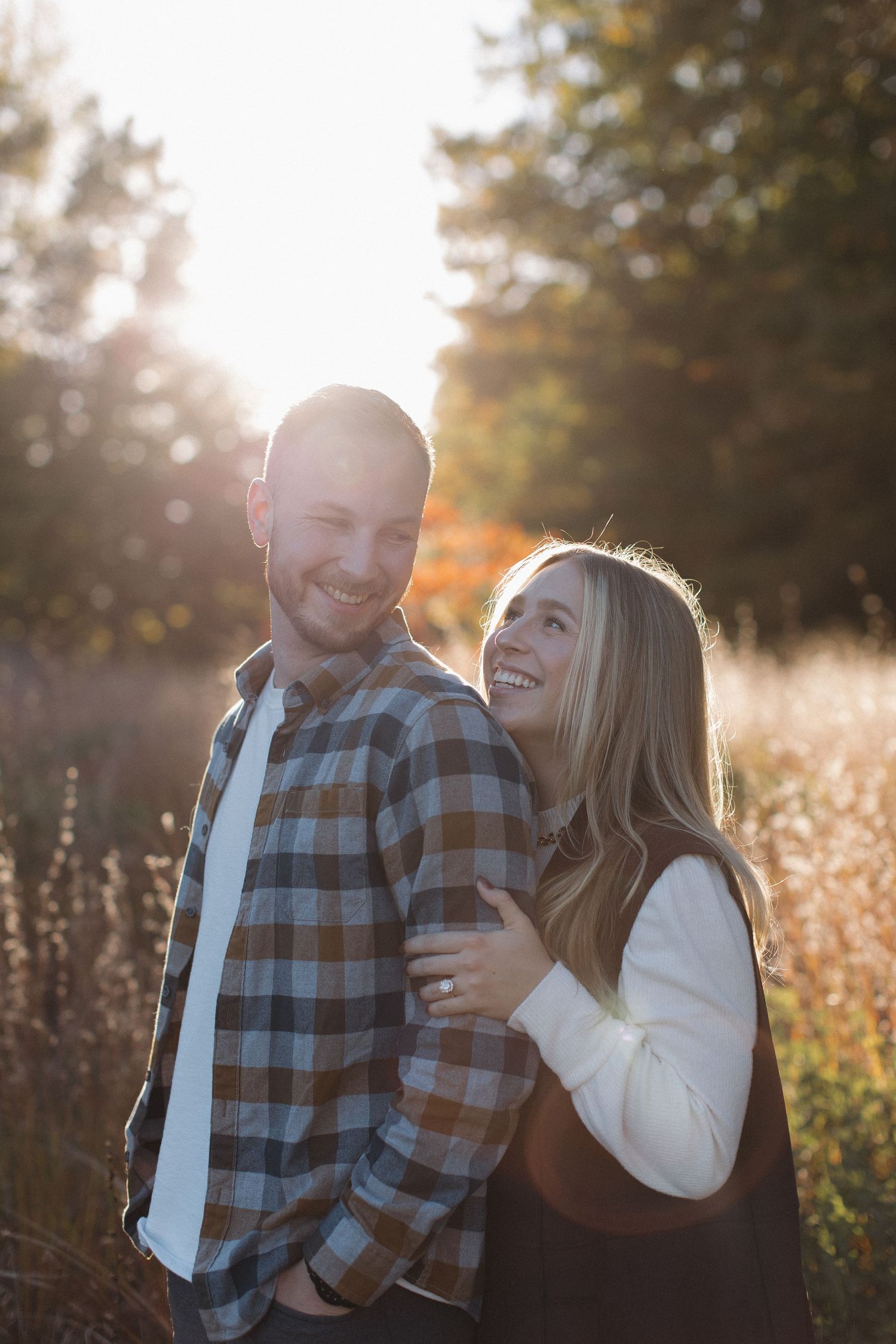 A couple shares a romantic moment in a sunlit field with fall foliage in the background.