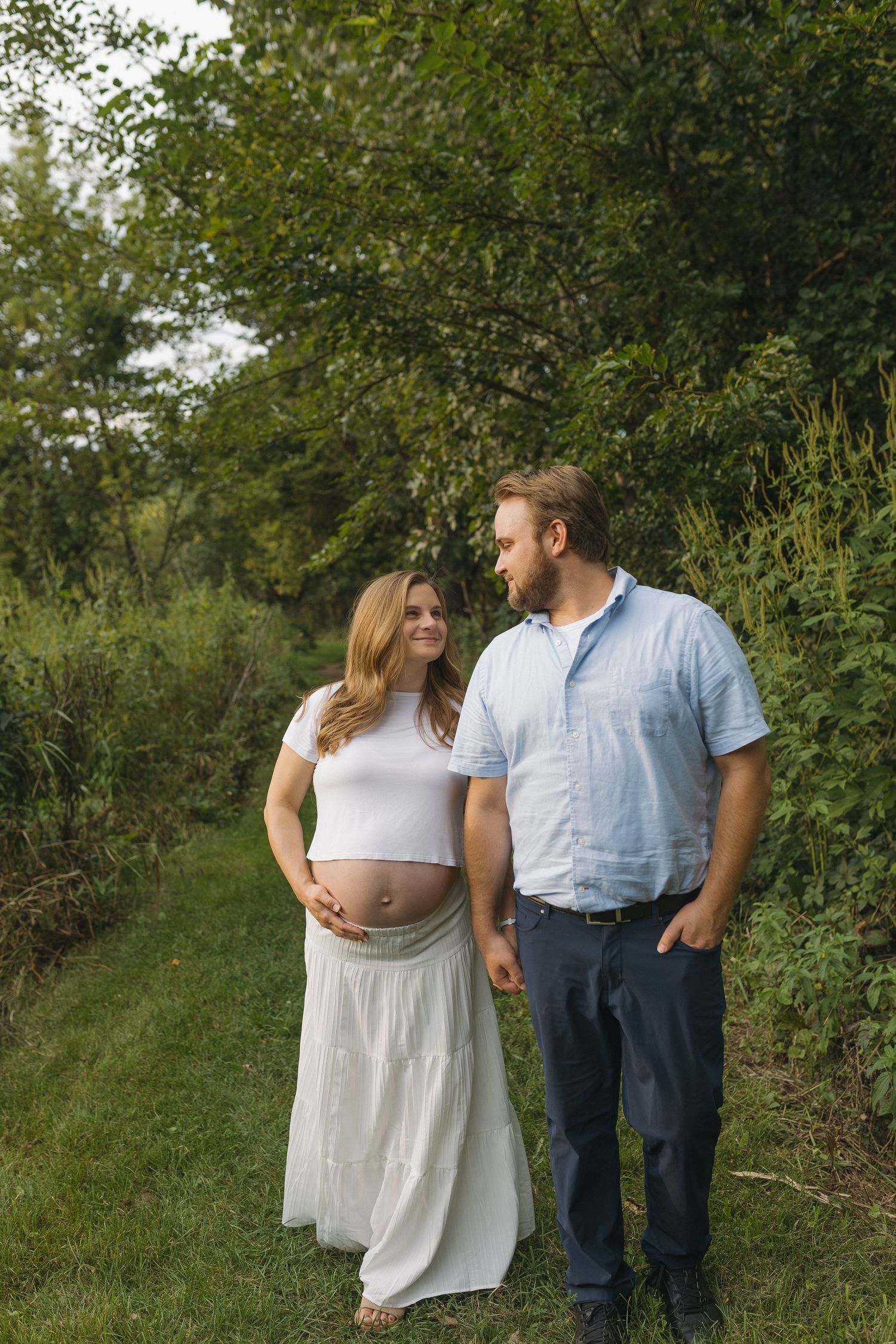 Pregnant woman in white dress holds baby bump while couple smiles at each other outdoors in lush green setting.
