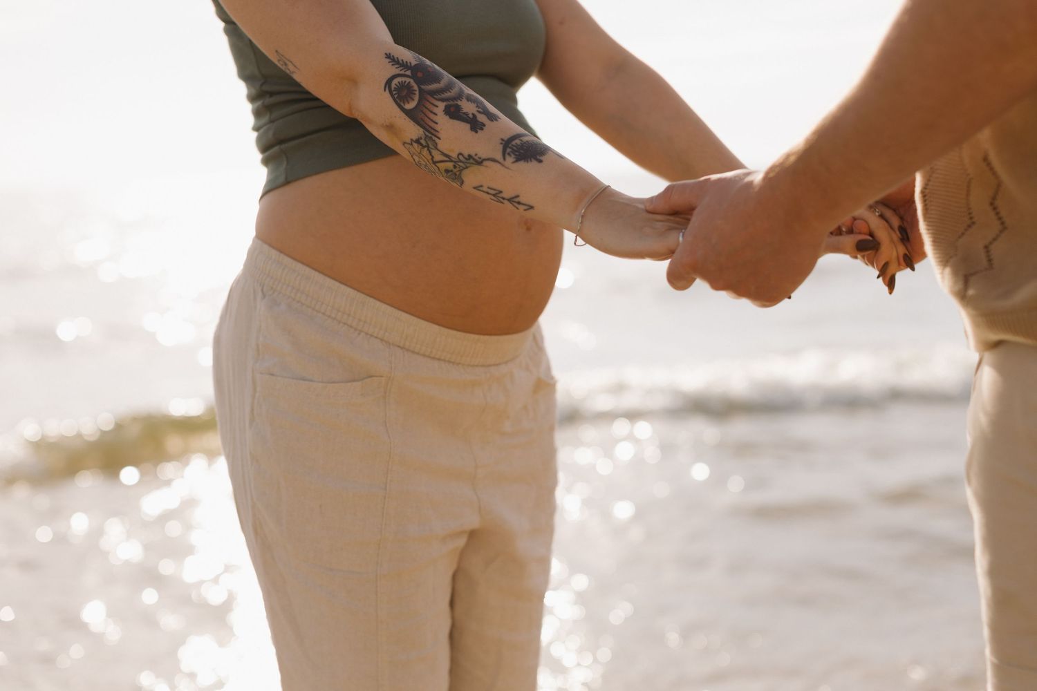 Pregnant woman with tattoos holding hands with partner on sunny beach, showing baby bump in crop top and linen pants.