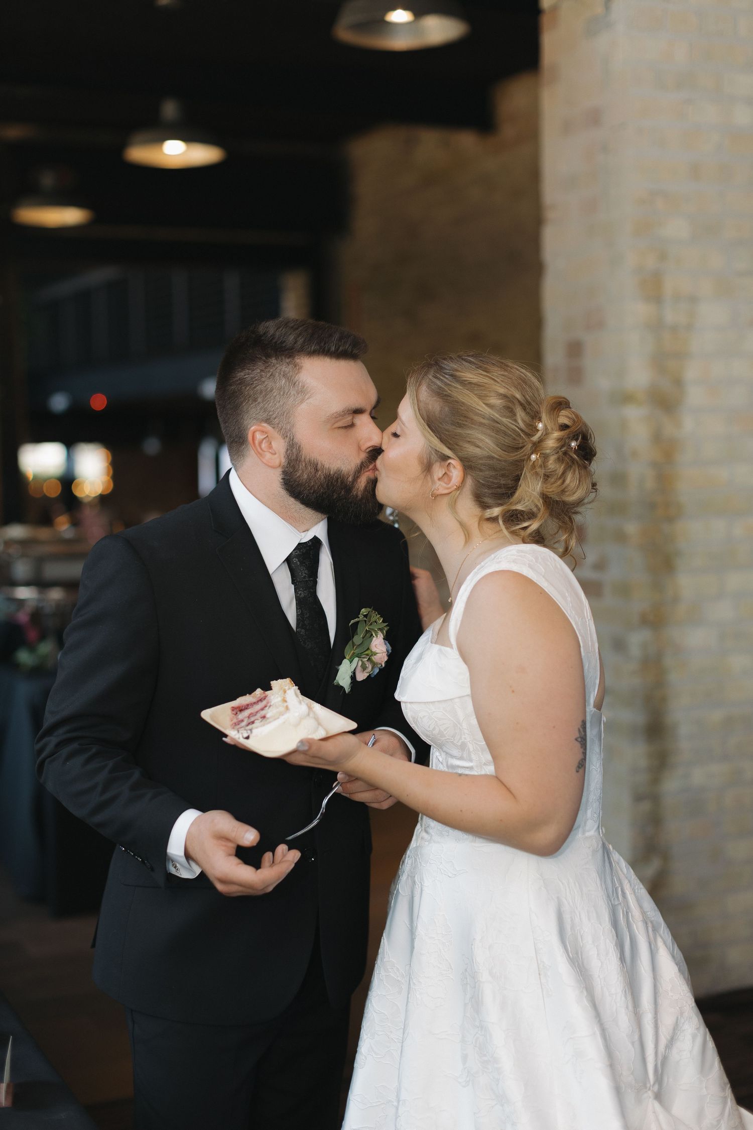 Bride and groom share a kiss while holding wedding cake at their indoor reception venue.