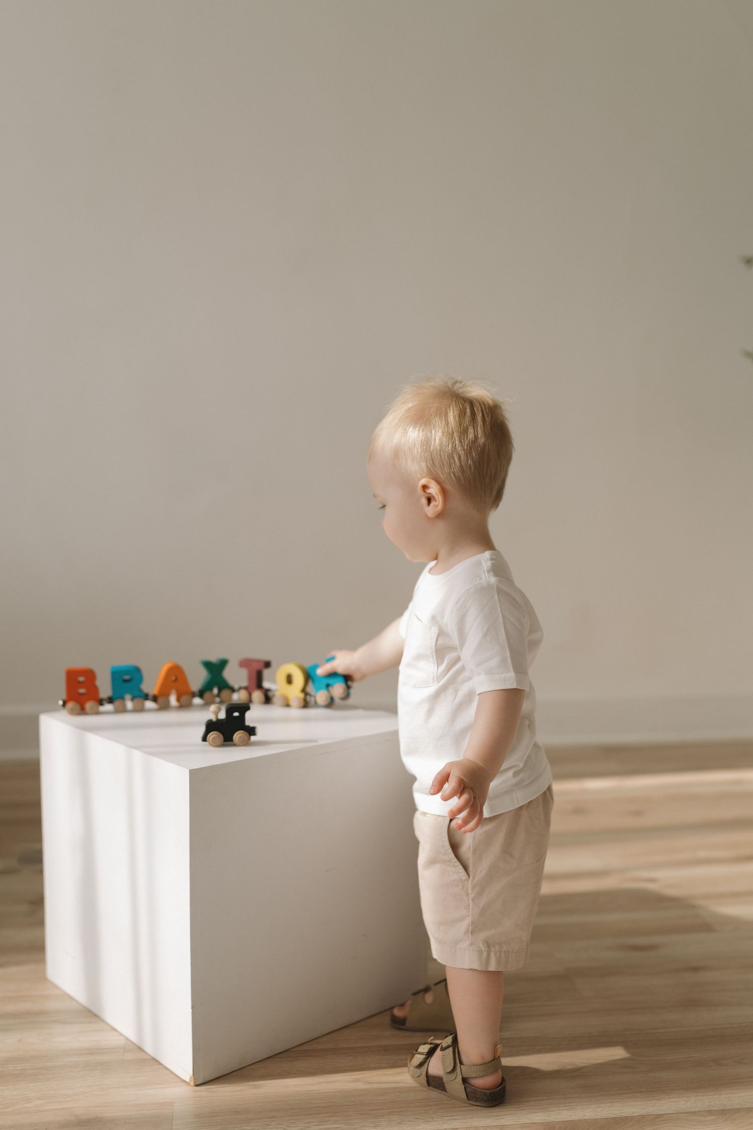 Toddler boy in white shirt playing with colorful letter toys spelling a name on a white cube table.
