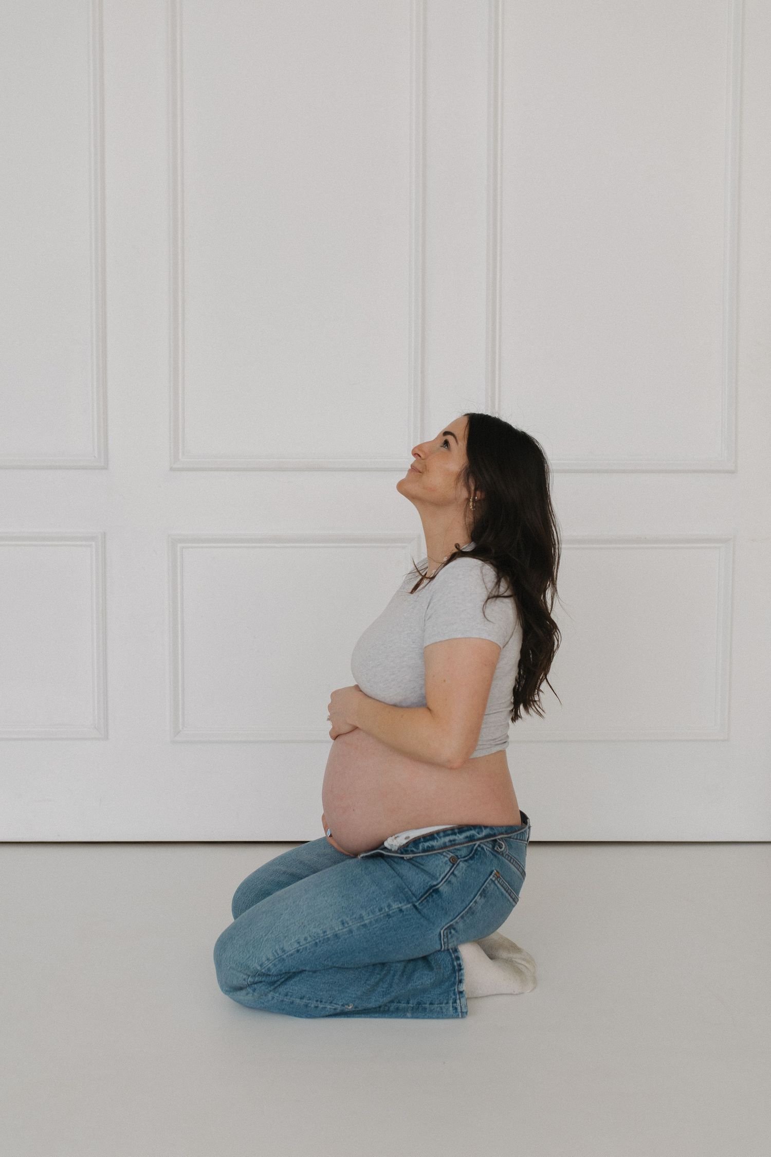 Side view of a pregnant person sitting against a white paneled wall.