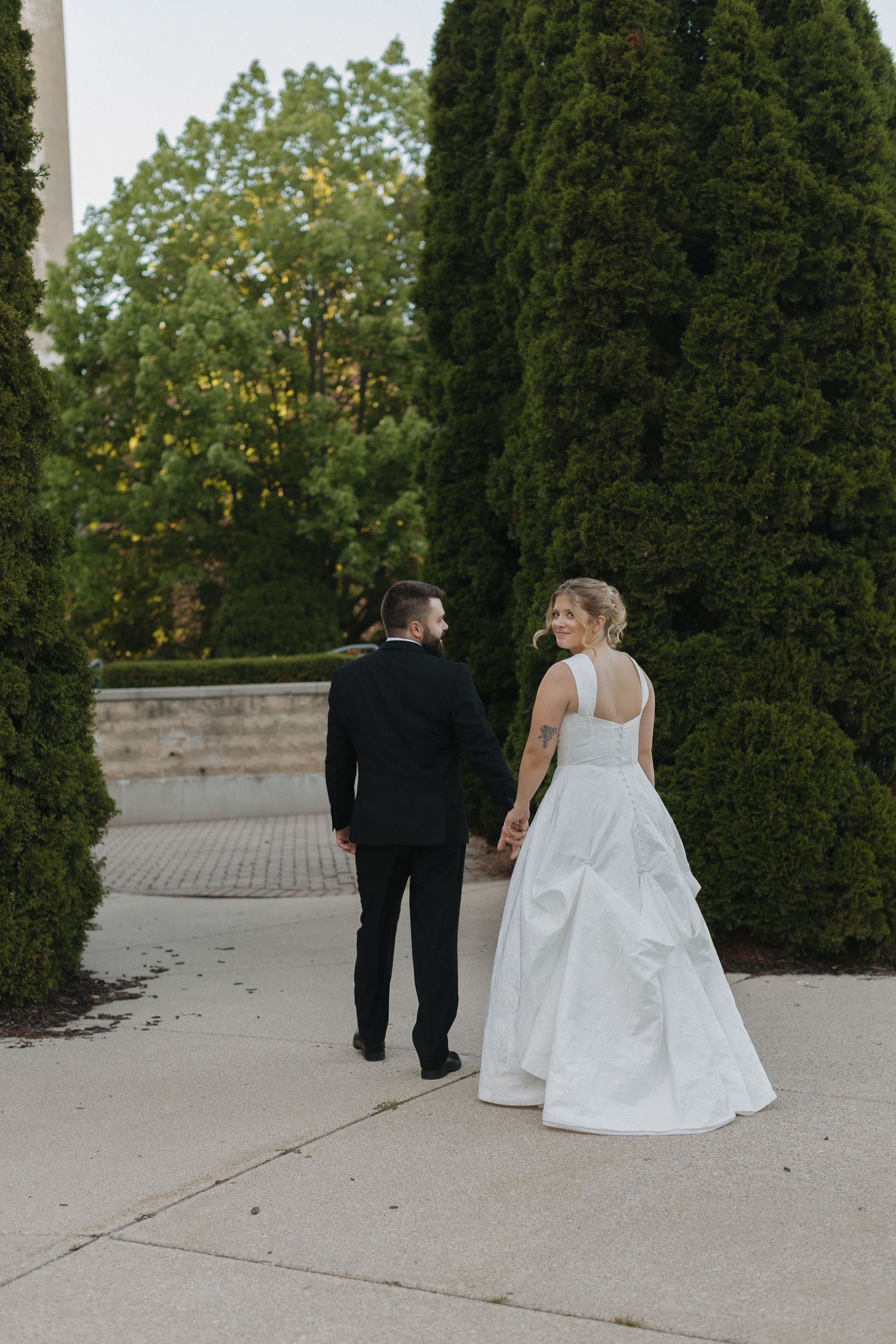 Bride and groom walking hand in hand through tree-lined path, bride glancing back in elegant white gown.
