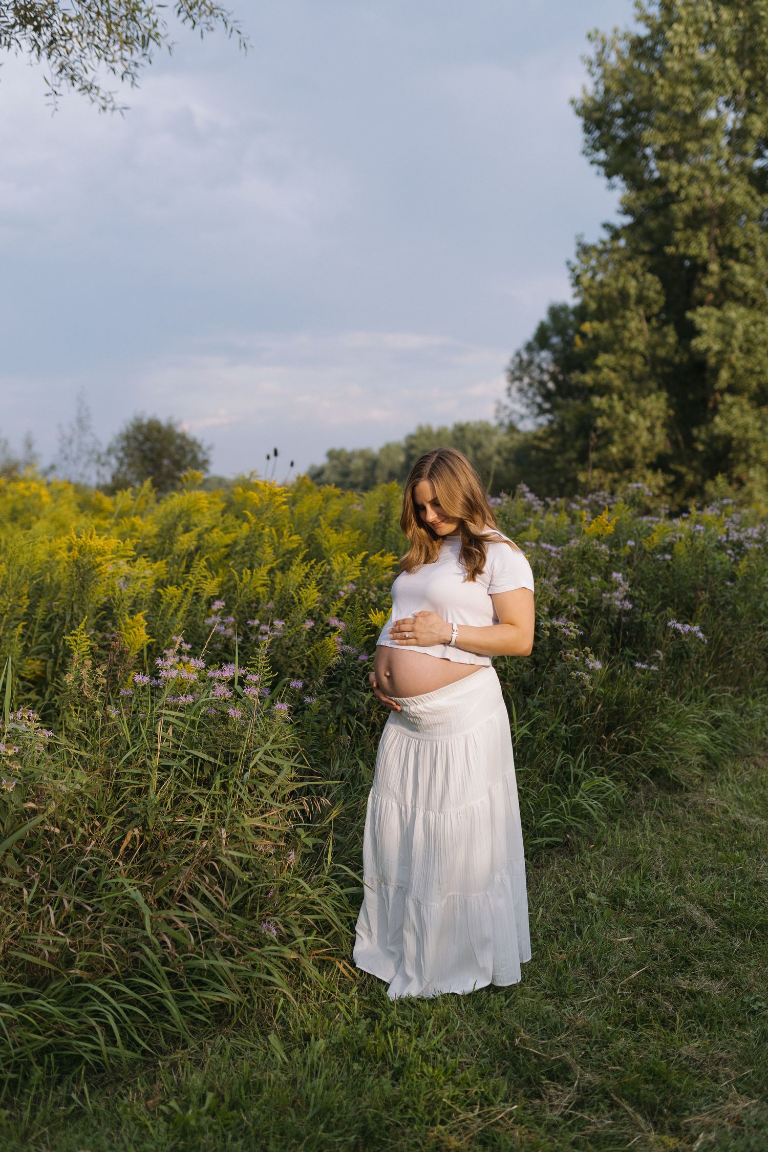 Pregnant woman in white dress cradling baby bump in a wildflower meadow with golden blooms and green trees.