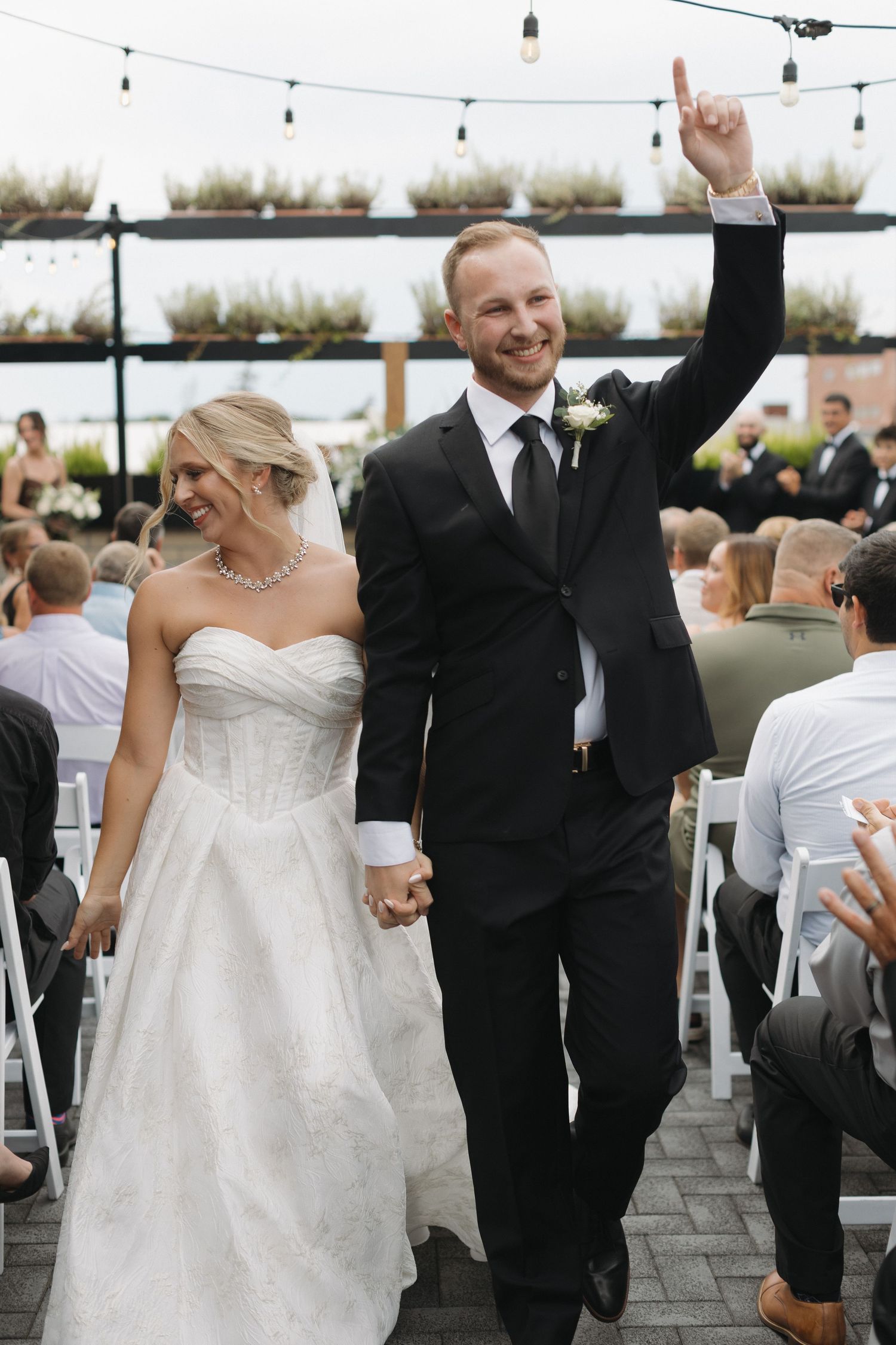 Joyful newlyweds walk down the aisle, groom raising his hand in celebration, bride in a strapless white gown.