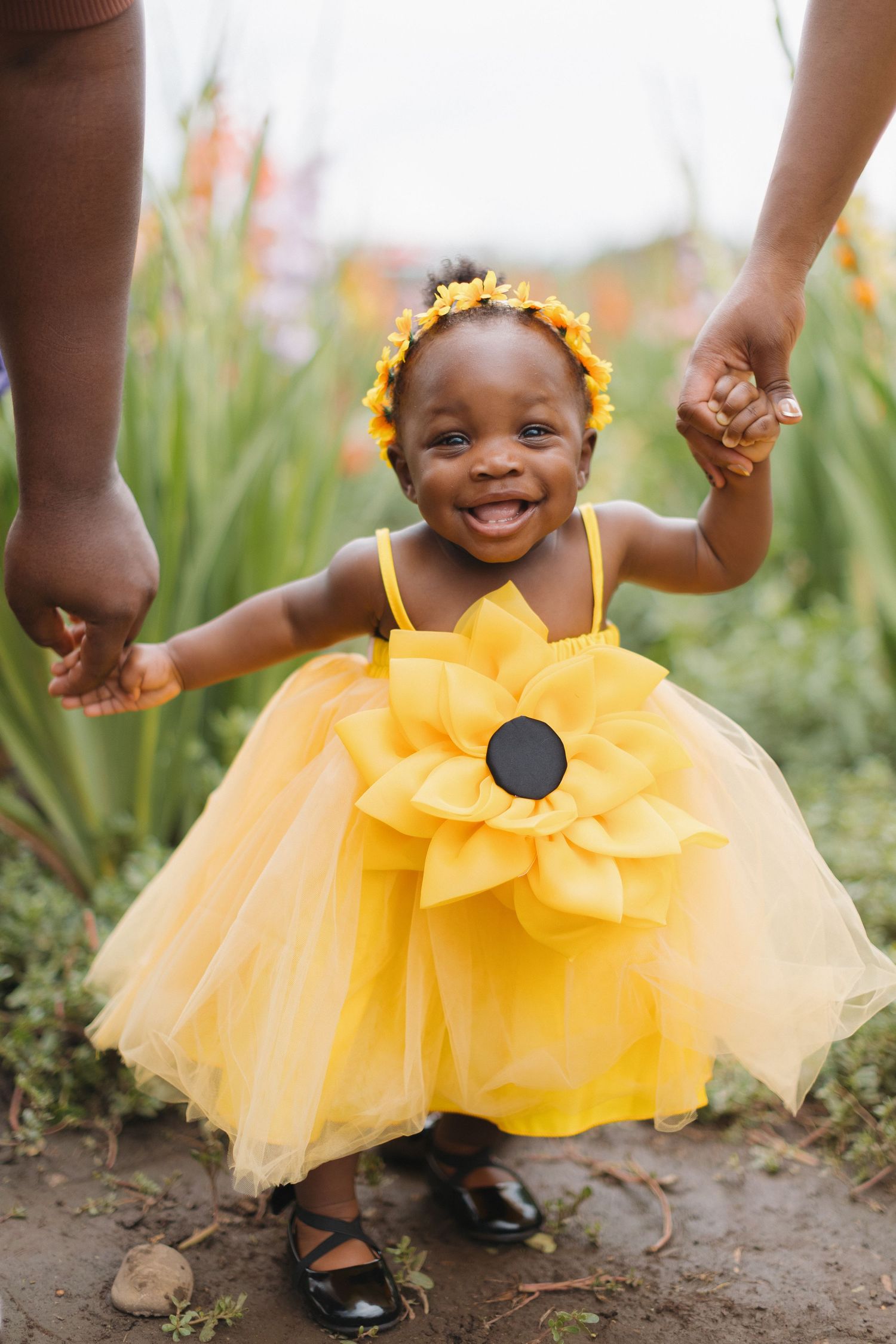 A joyful baby girl in a yellow sunflower tutu dress and floral headband holds hands with adults in a garden.