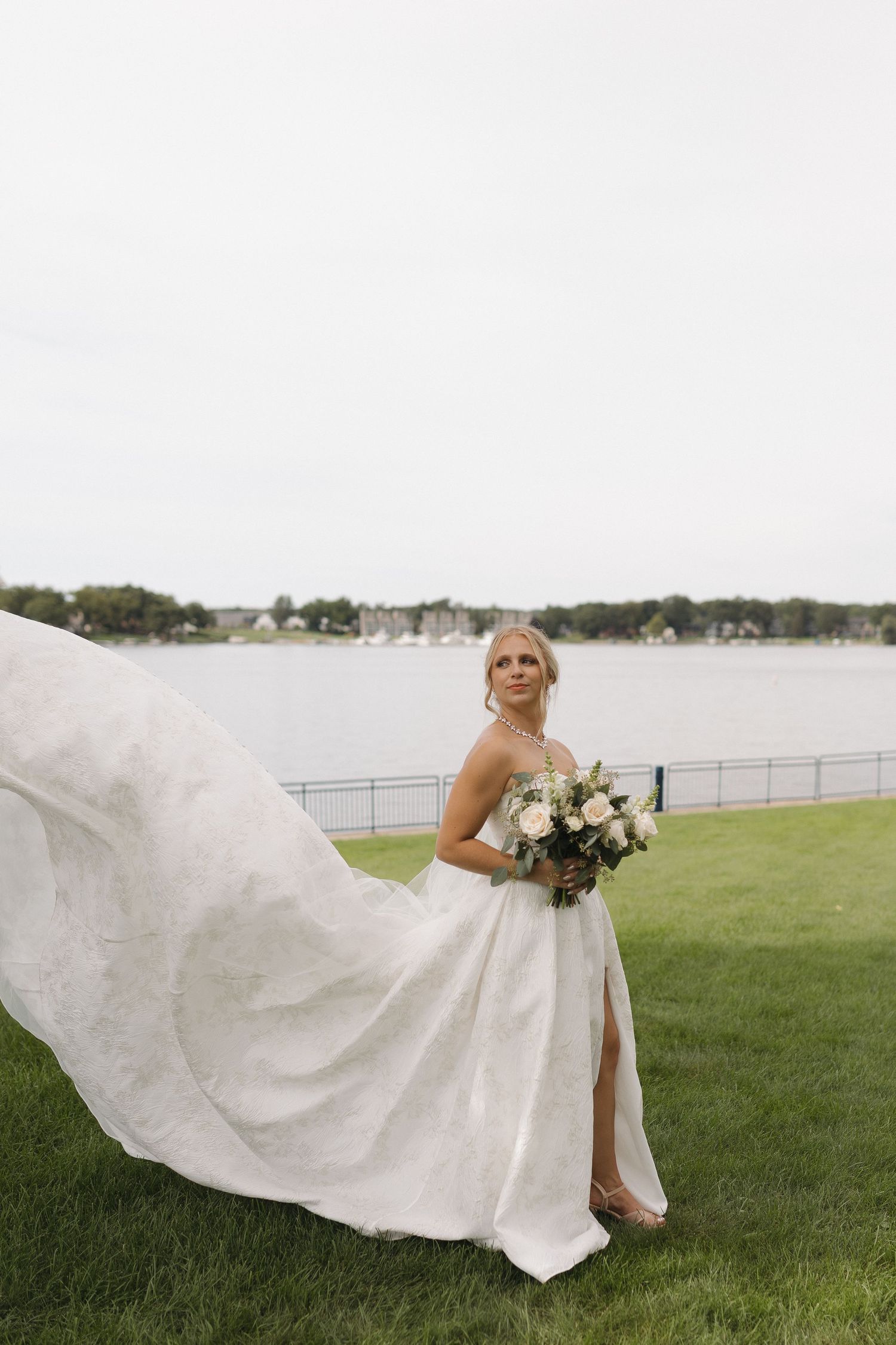 Bride in flowing white wedding gown holds bouquet by lakeside on green lawn, dress billowing in the breeze.