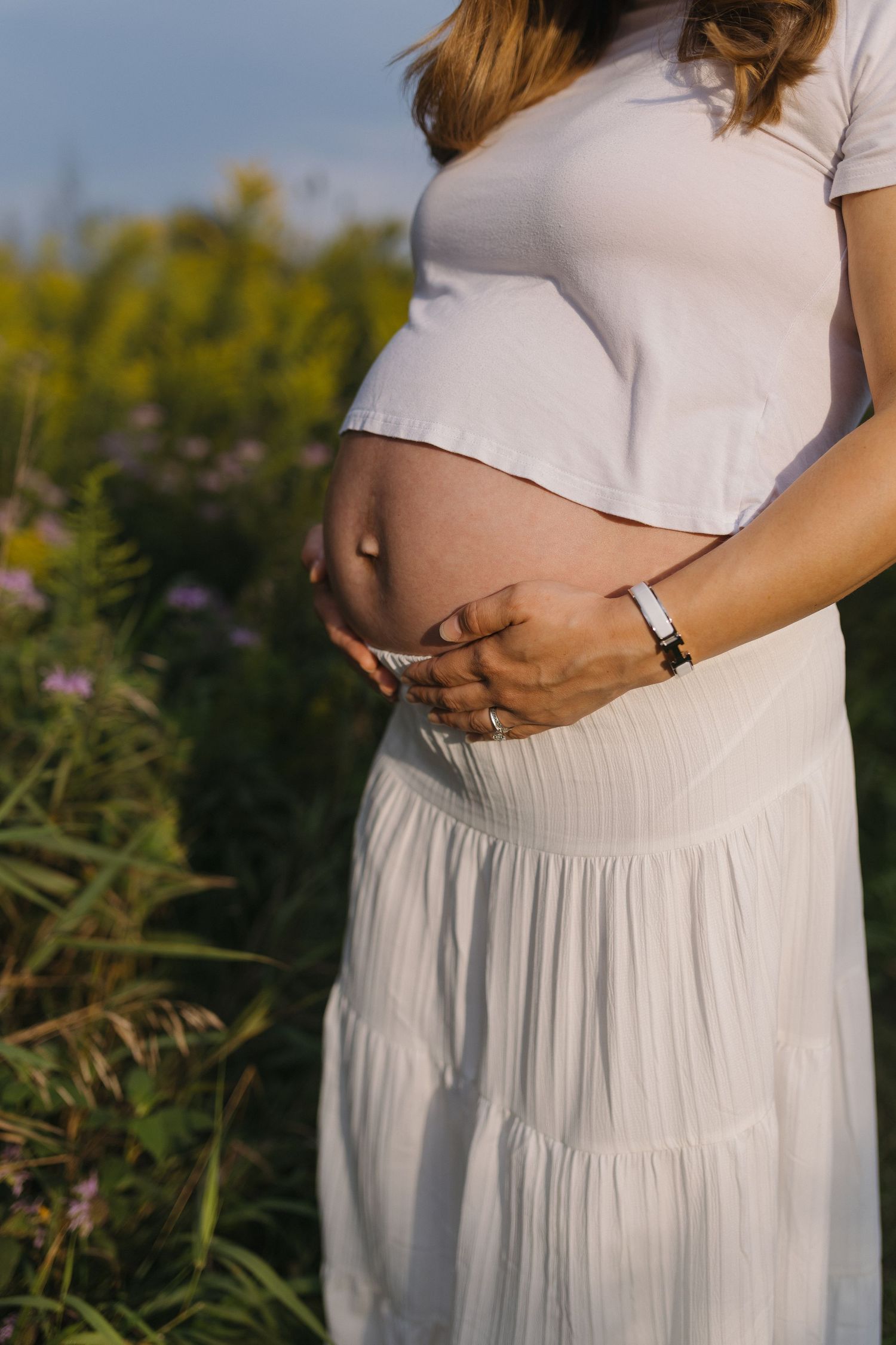 Pregnant woman in white crop top and skirt cradling baby bump outdoors in a sunny flower field.