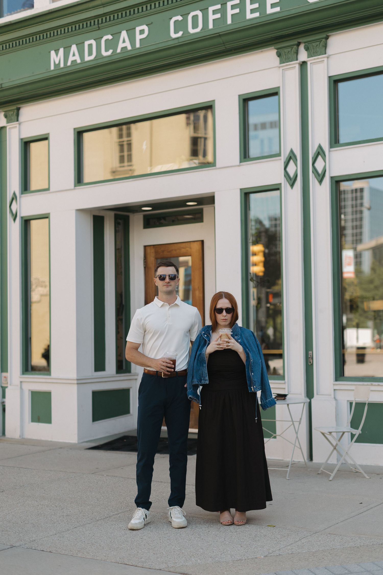 Couple standing outside Madcap Coffee shop, man in white polo and woman in black dress with denim jacket, both wearing sunglasses.