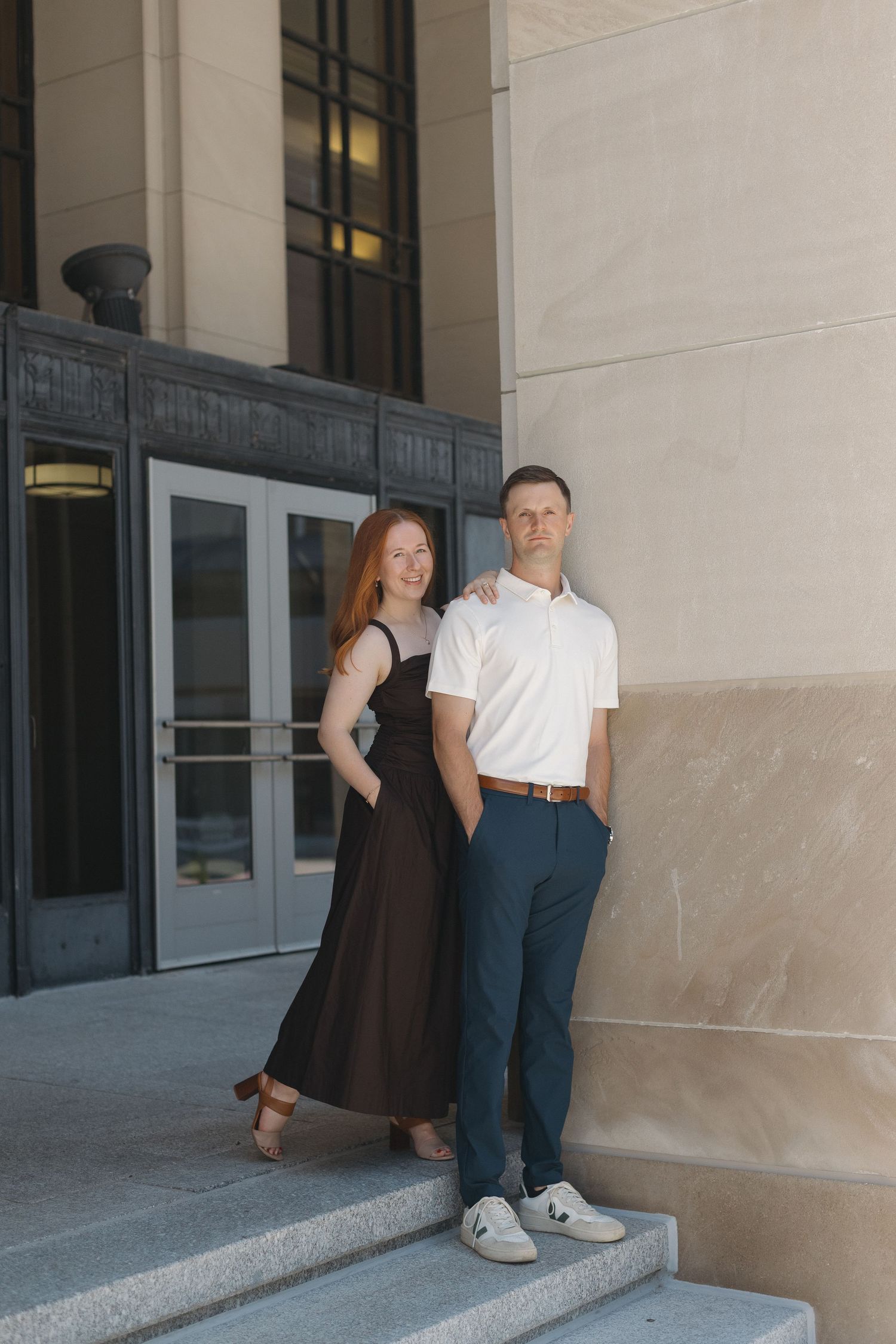 A couple poses on steps outside a building, woman in black dress and man in white polo and navy pants.