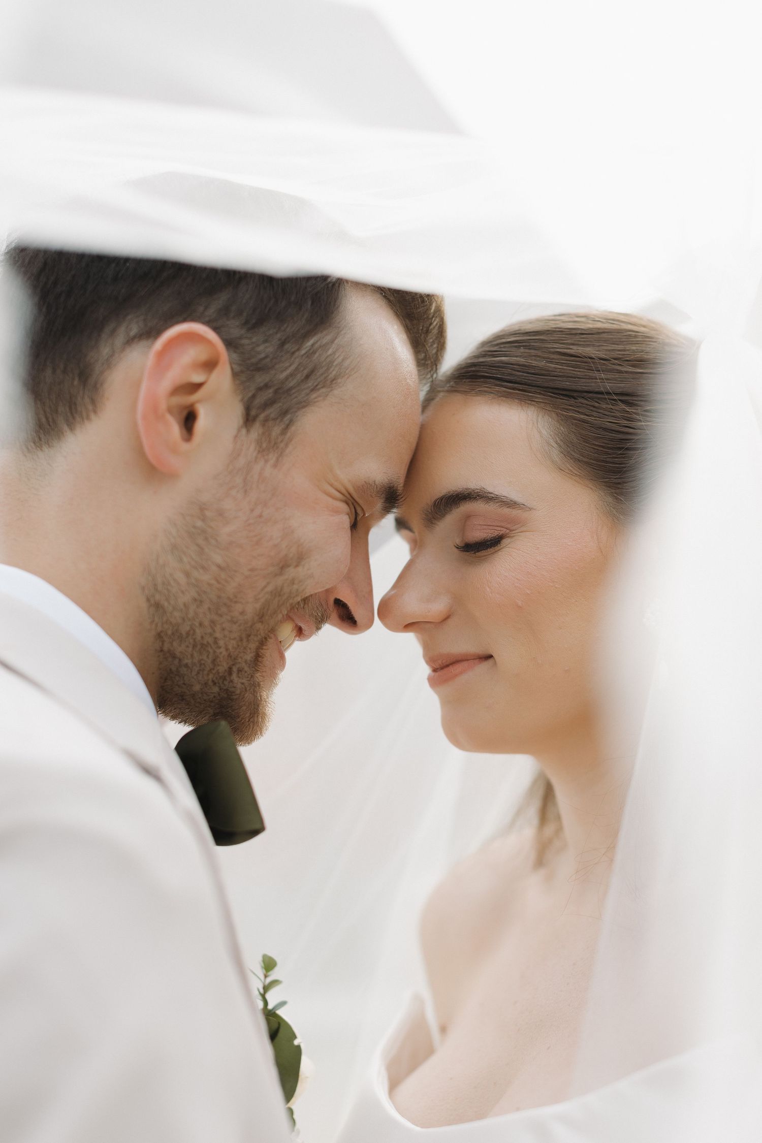 A bride and groom touch foreheads tenderly under a flowing white veil on their wedding day.
