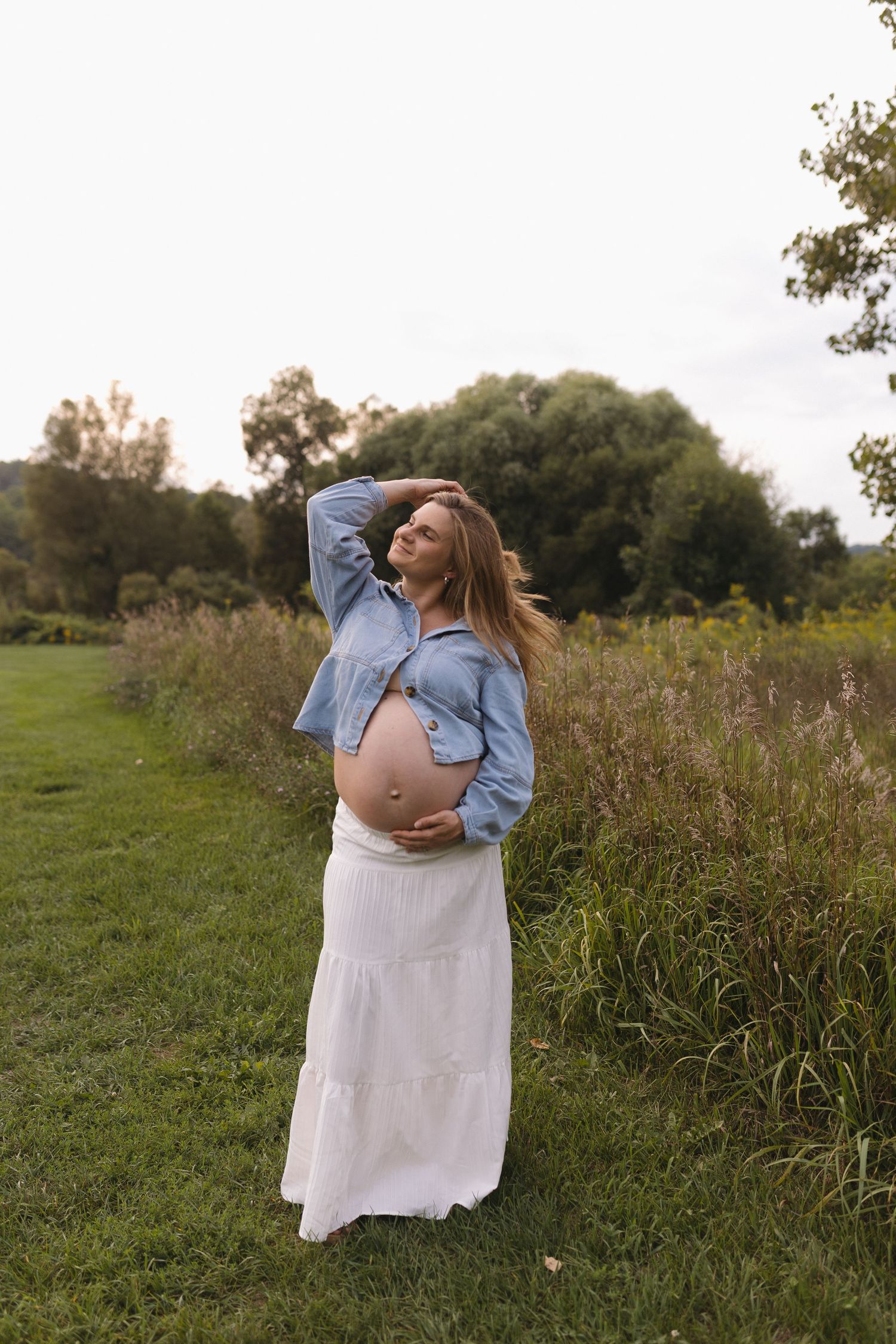 Pregnant woman in denim jacket and white skirt cradling her baby bump outdoors in a lush green field.