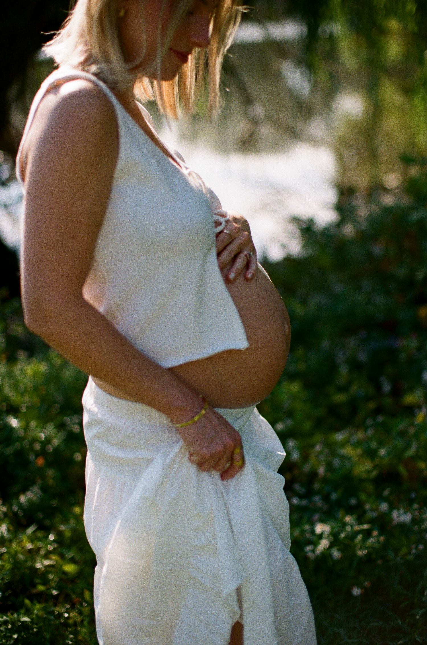 Pregnant woman in white tank top and skirt cradling her baby bump outdoors in a sunlit garden.