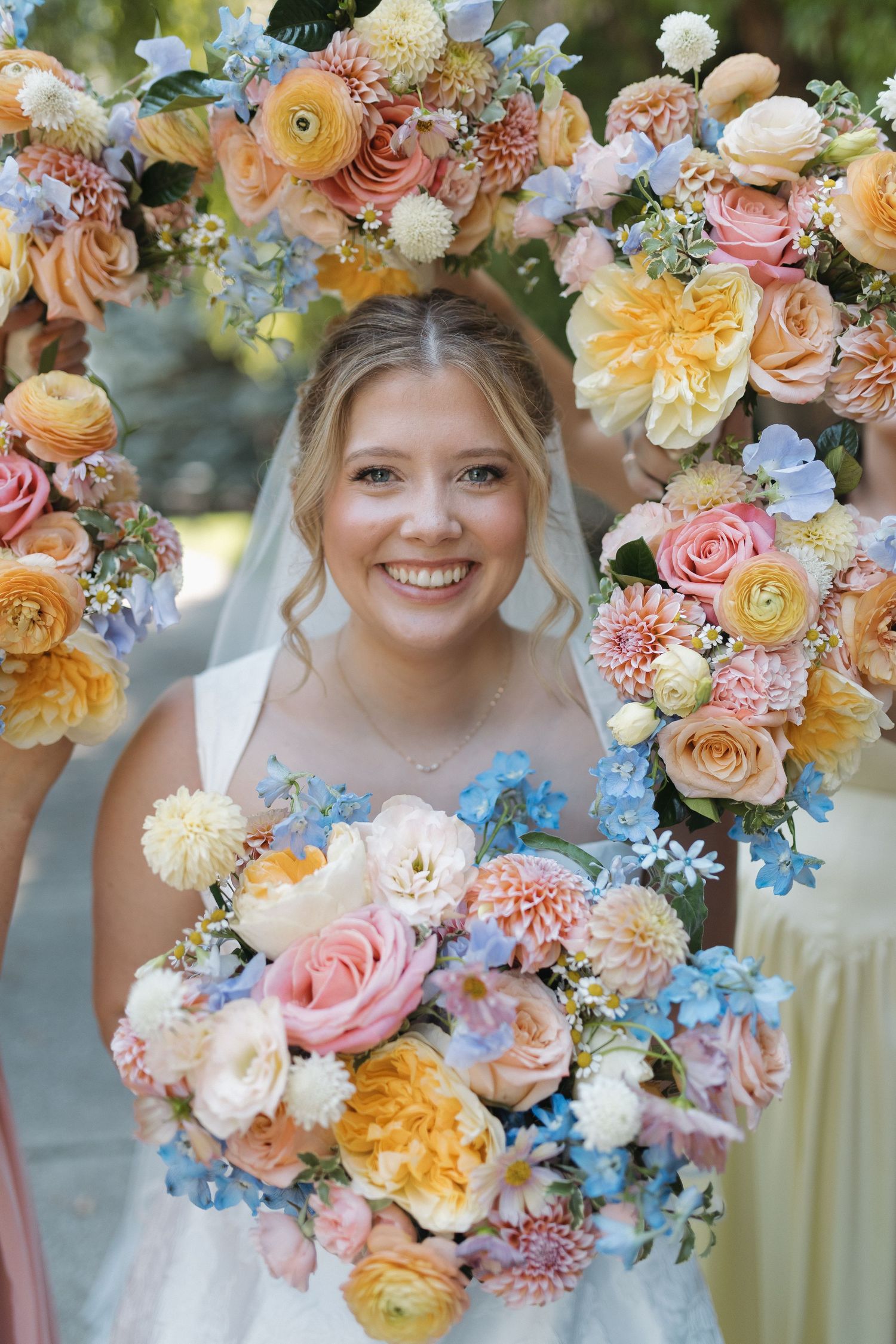Smiling bride in white dress and veil framed by colorful floral bouquets with pink, peach, and blue flowers.