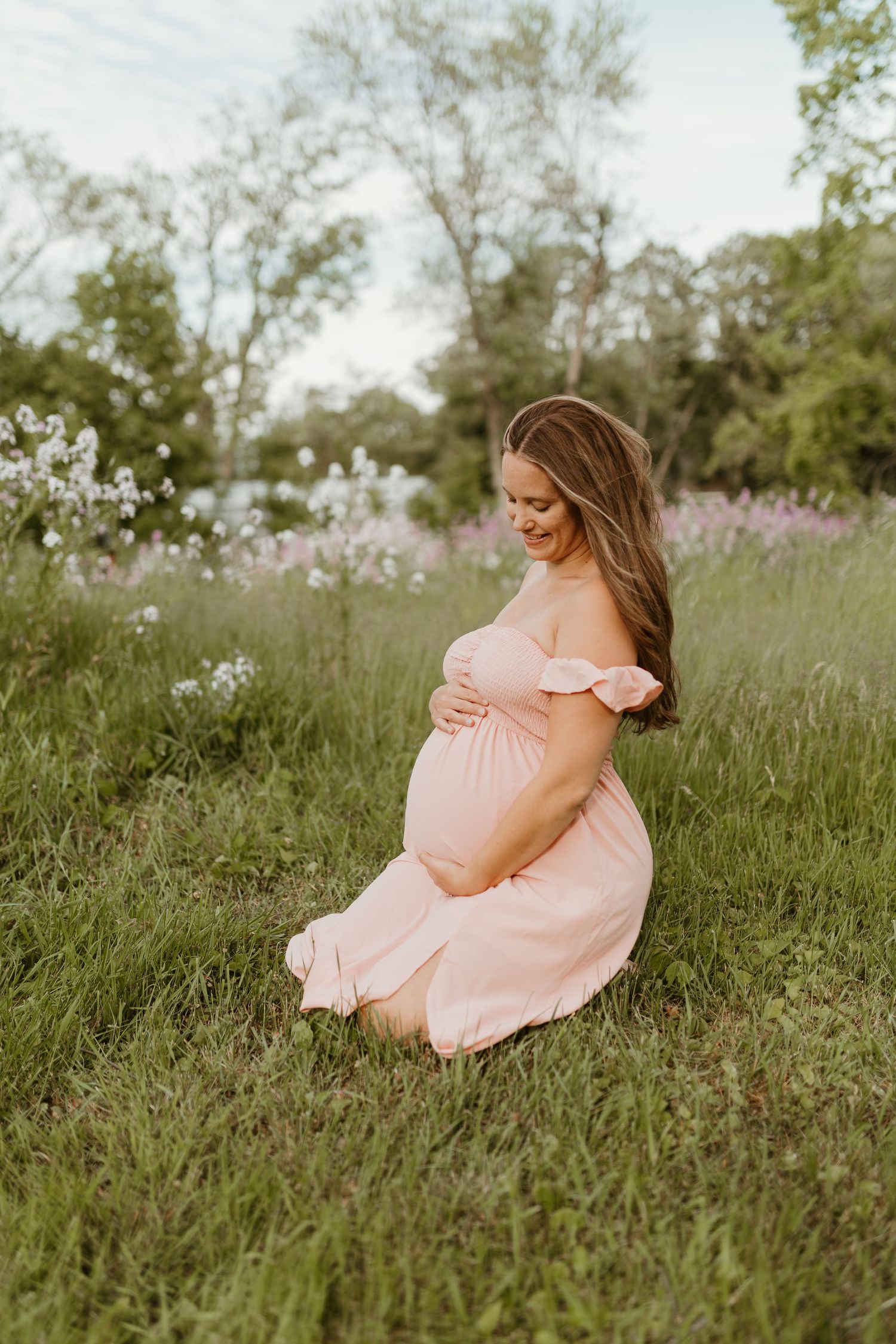A person in a peach-colored dress sits gracefully in a grassy field with blooming flowers.