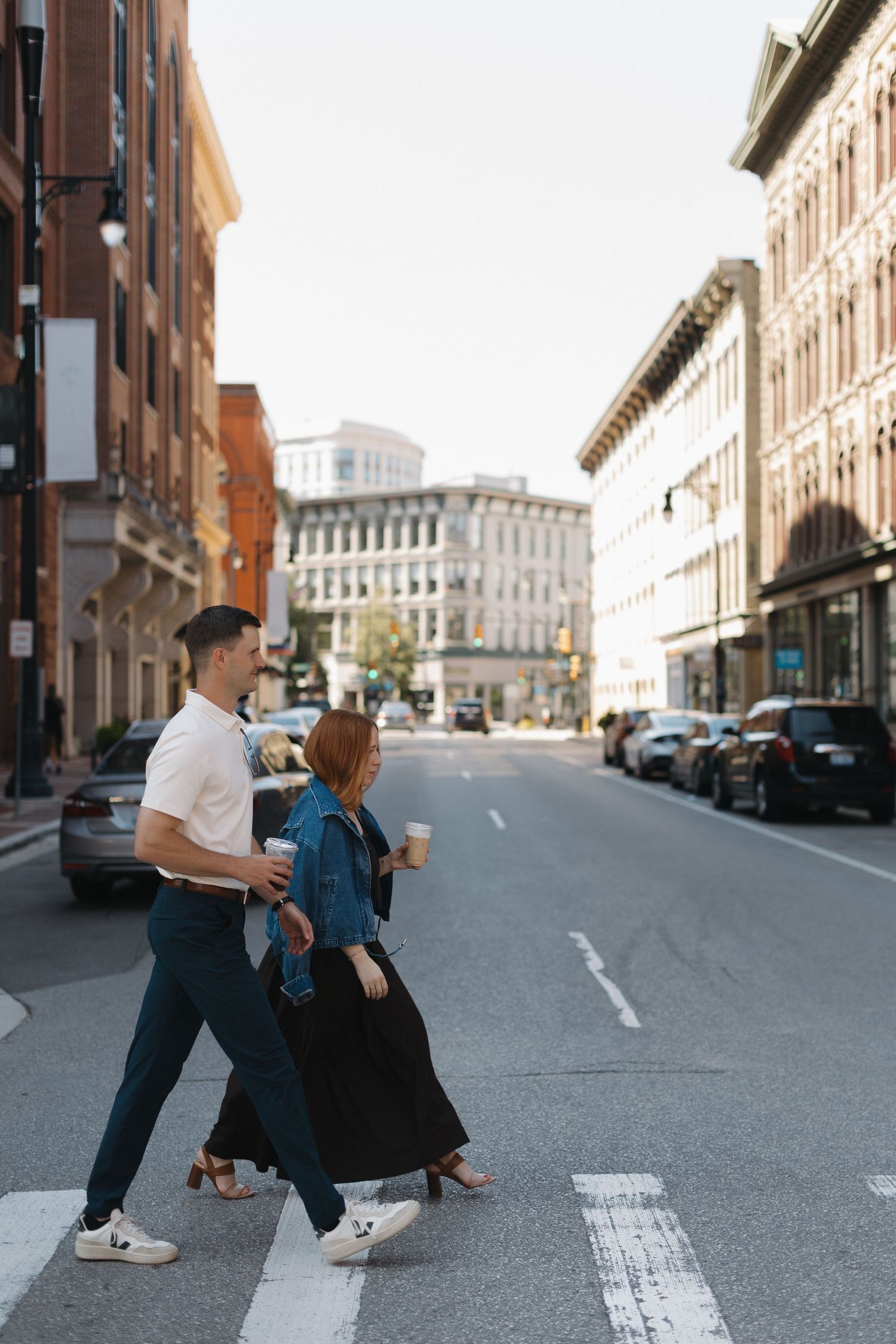 Couple crossing a city street crosswalk holding coffee cups, surrounded by historic brick buildings on a sunny day.