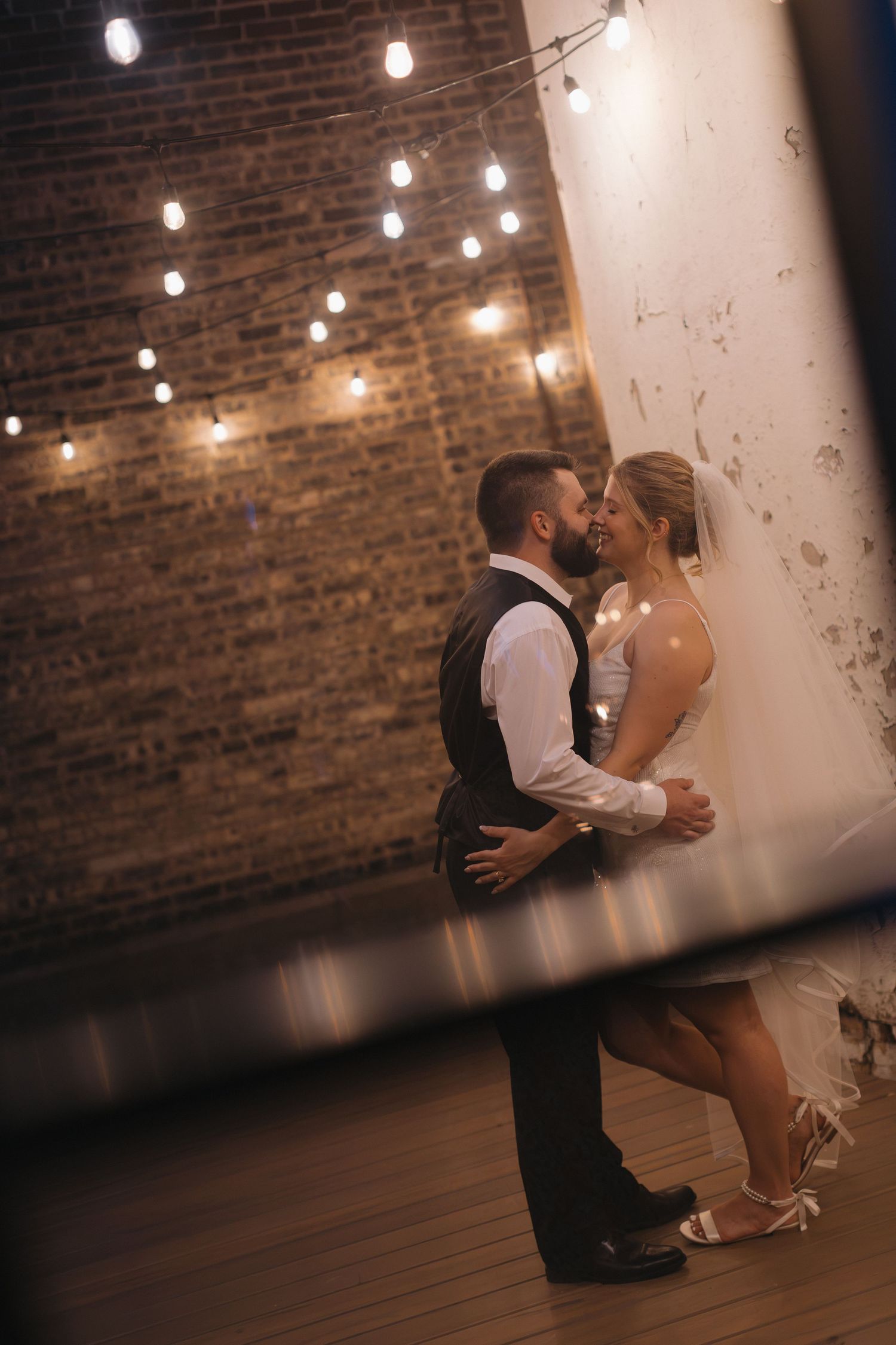 Bride and groom share a romantic first dance in a warmly lit venue with exposed brick walls and string lights.
