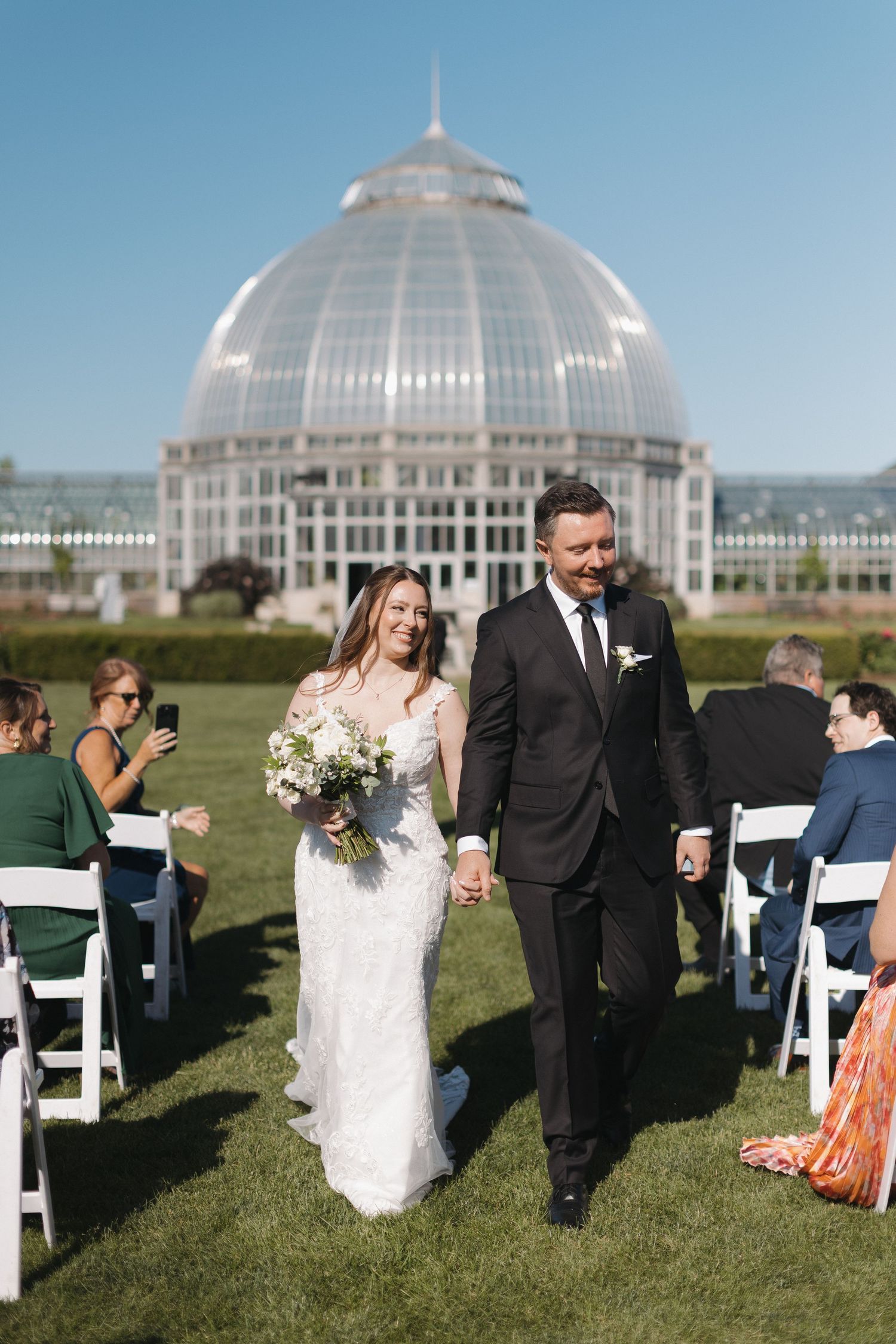 Bride and groom walk down outdoor aisle holding hands at Belle Isle Conservatory wedding ceremony on sunny day.