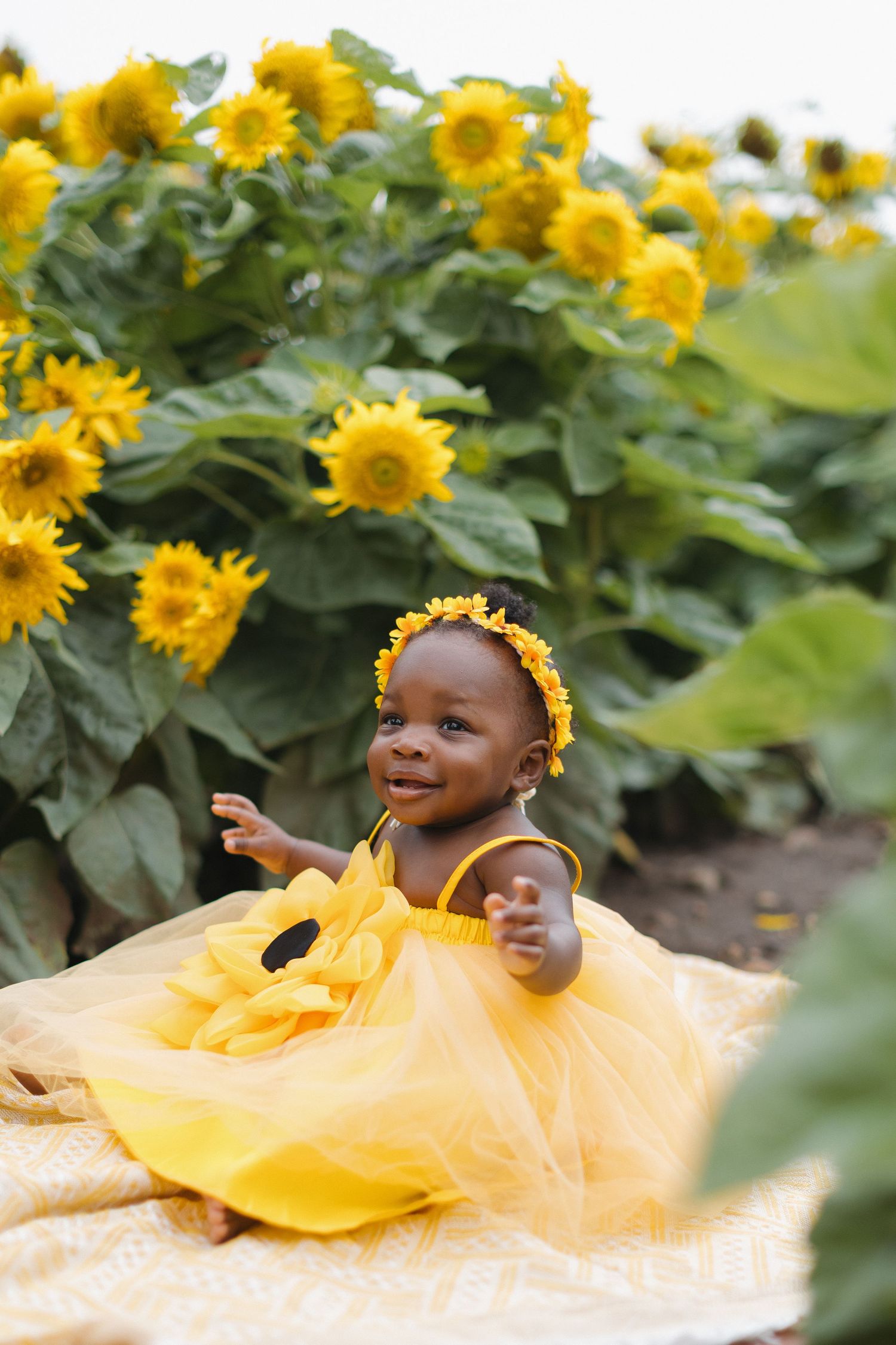 Adorable baby girl in yellow tutu dress and floral headband sitting in a sunflower field.