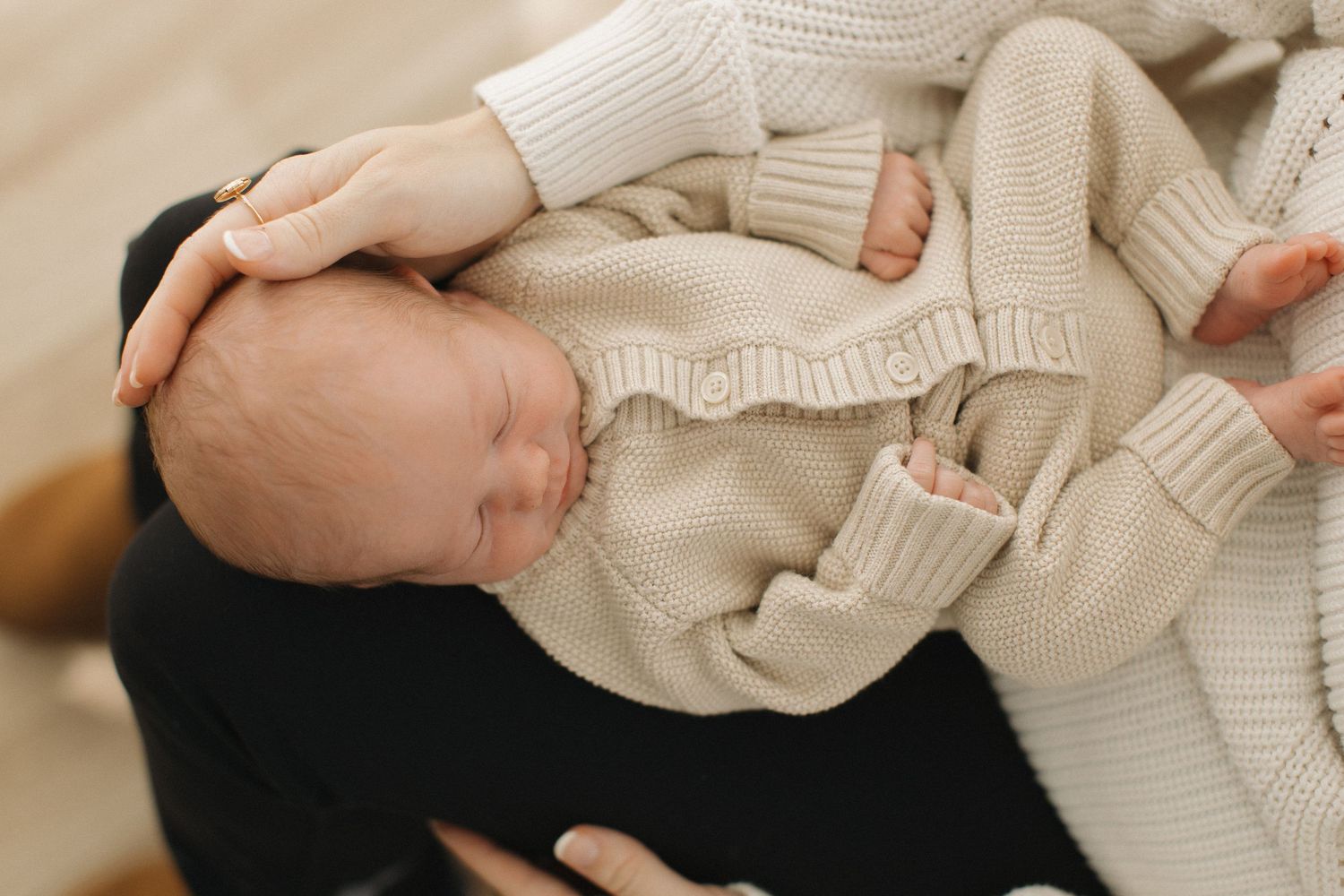 Newborn baby sleeping peacefully in cream knit outfit, cradled in parent's arms.