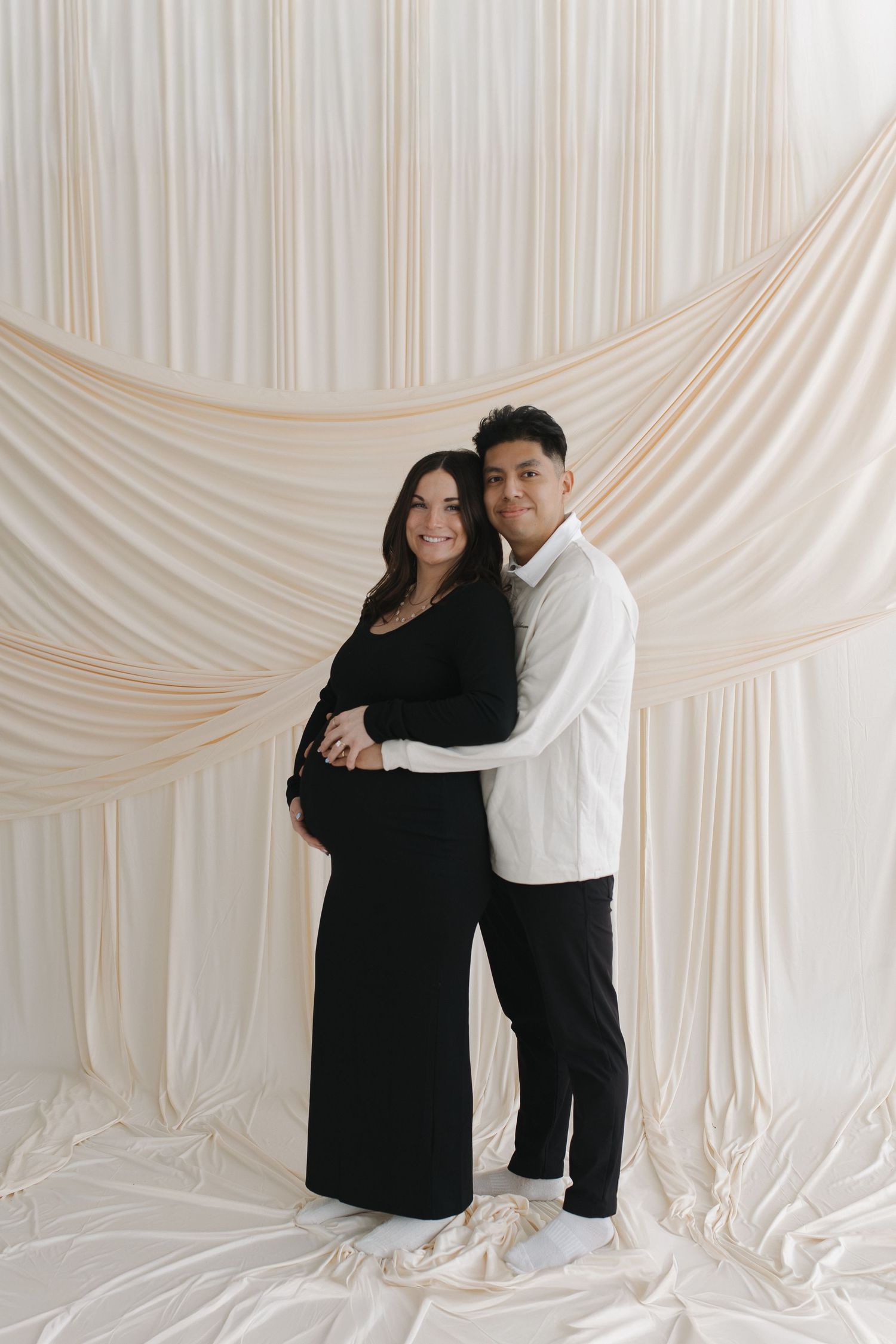 A couple embraces tenderly against a draped white fabric backdrop in a studio setting.