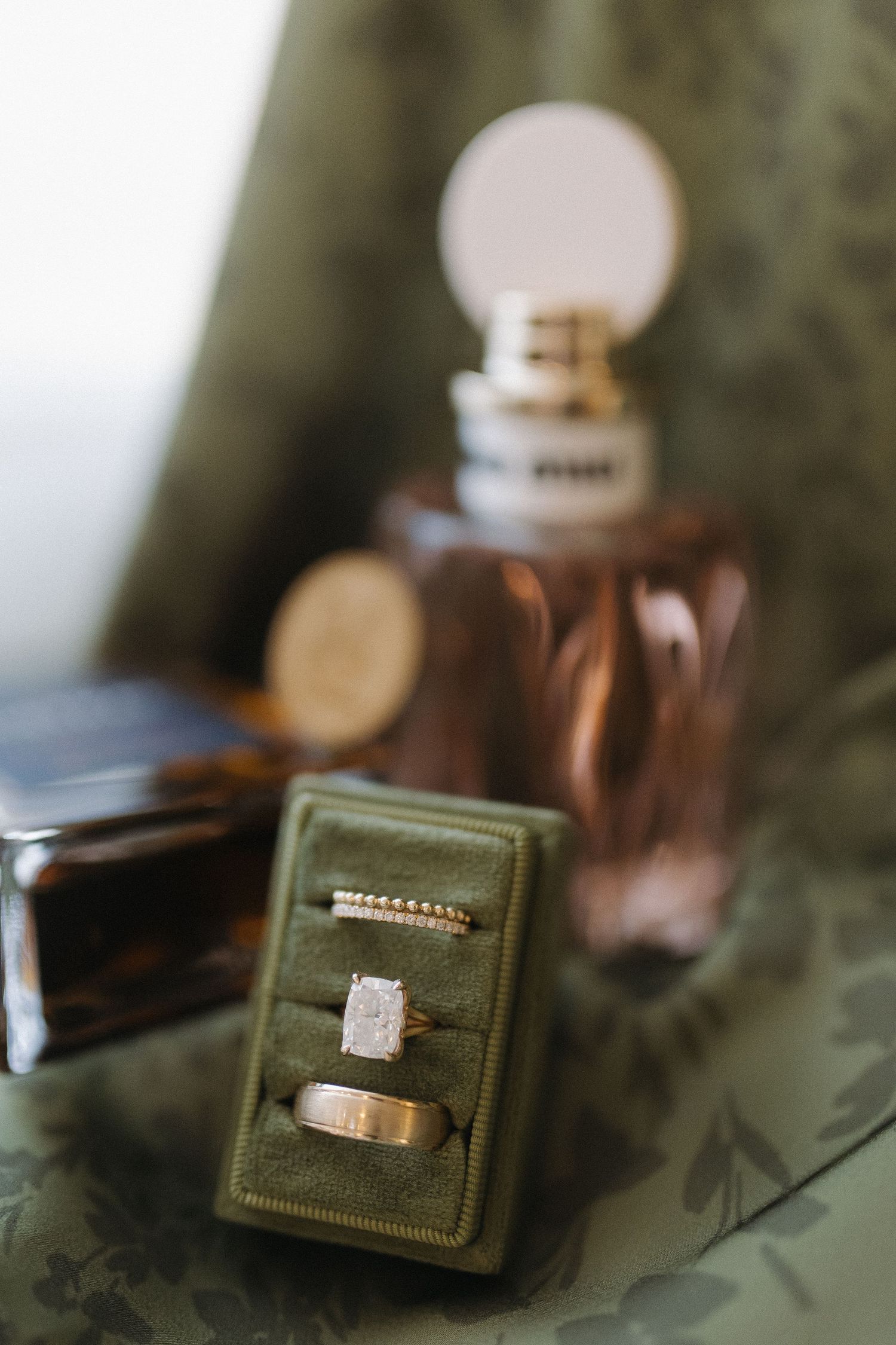 Three wedding rings displayed in a green velvet ring box with perfume bottles in the background.