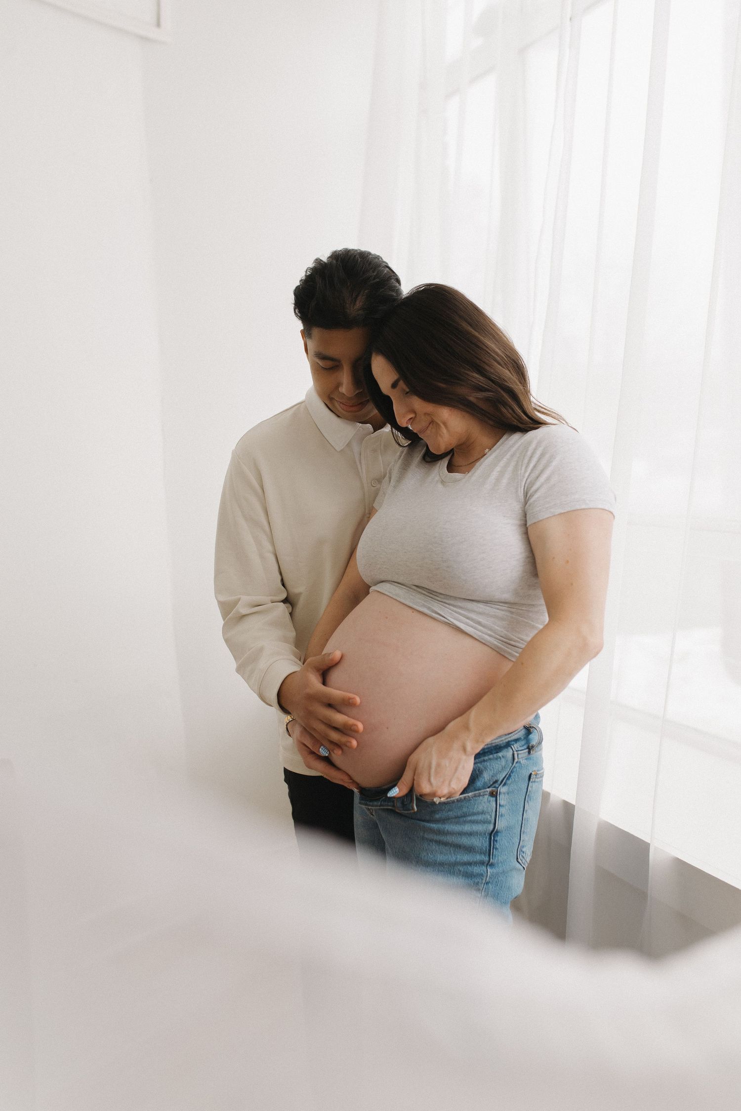 A couple tenderly cradles a pregnant belly during a maternity photo session in a bright, airy white room.