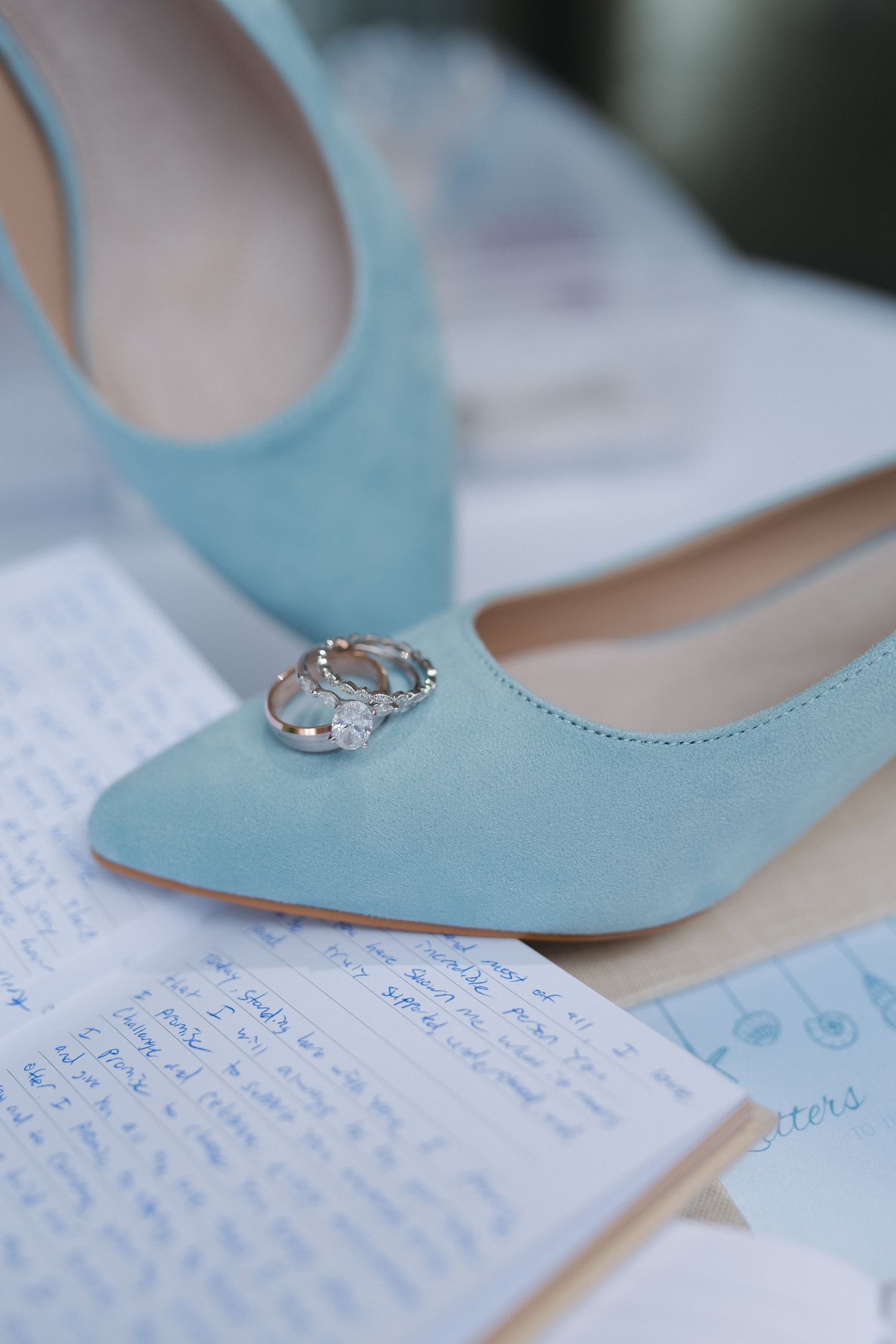 Light blue suede wedding shoes with diamond engagement and wedding rings resting on top, beside handwritten vows.