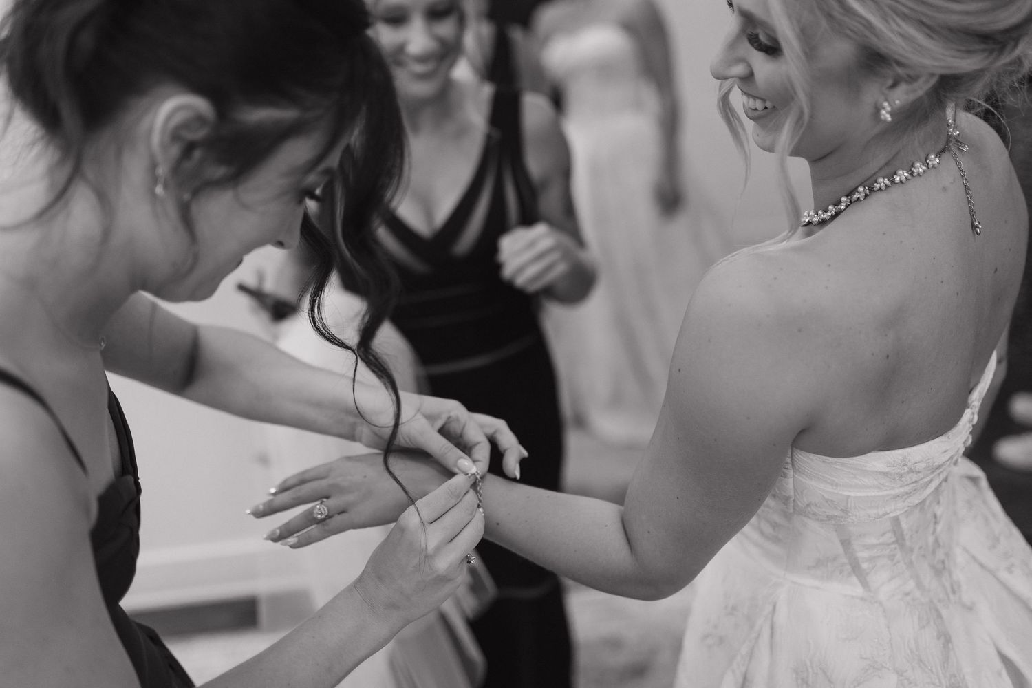 Bridesmaid helping bride with wedding dress details in black and white photo with guests in background.