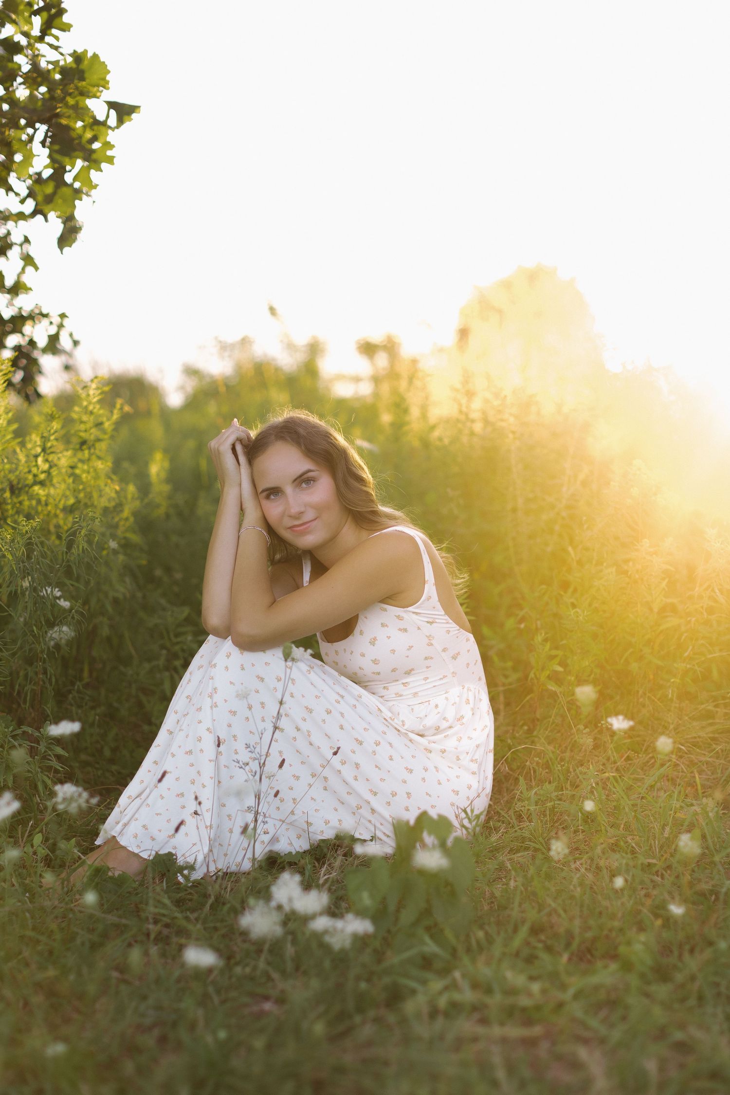 Young woman in white floral dress sitting in sunlit meadow with wildflowers, golden hour sunlight glowing behind her.