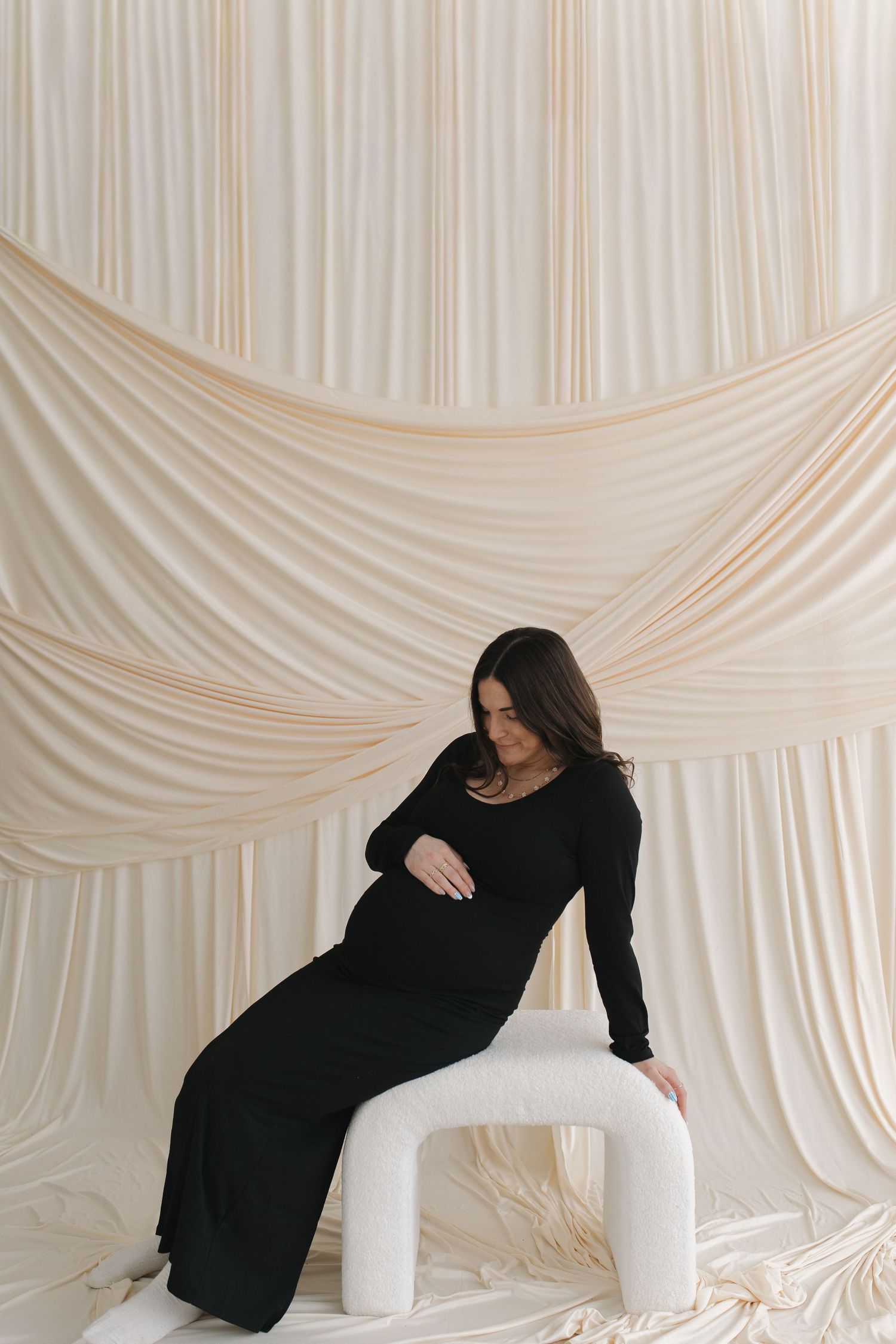 A person in a black dress reclines on white cushions against an elegant draped backdrop.