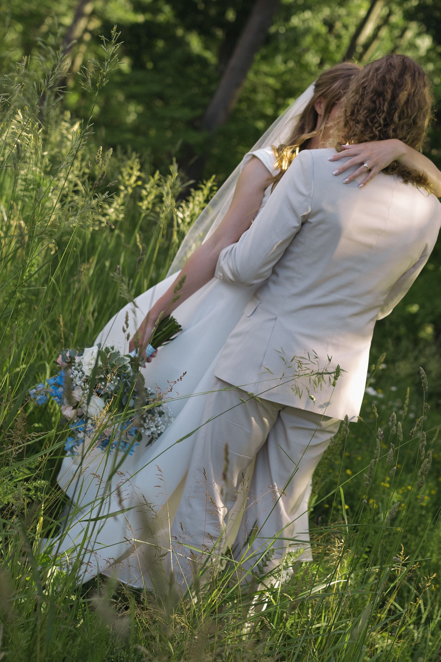 Bride and groom embracing in a lush green meadow, bride holding a blue and white bouquet, wearing a white gown and veil.
