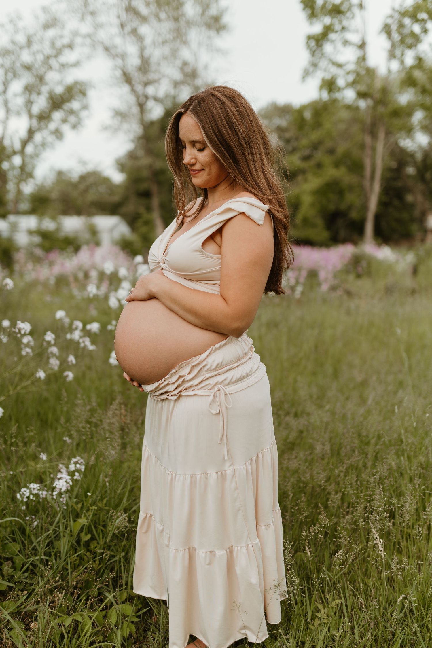 Maternity photos in flowing cream dress amid wildflower field at sunset.