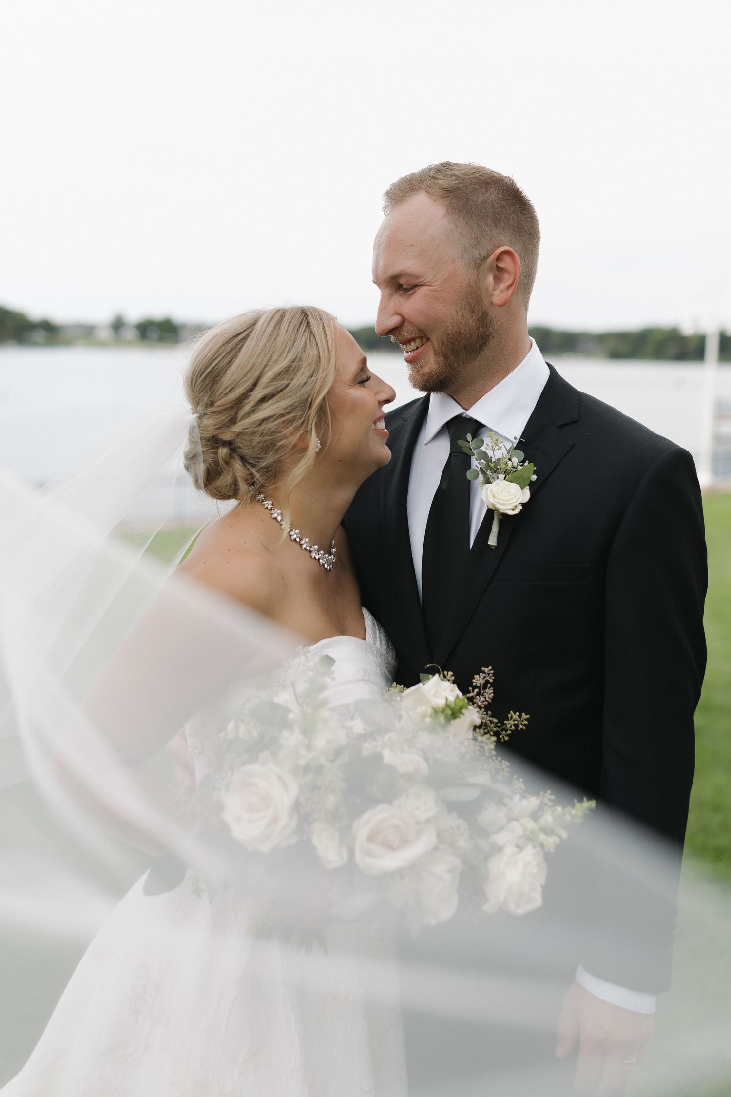 Bride and groom share a romantic moment outdoors near water, veil flowing, as groom wears black suit with white boutonniere.